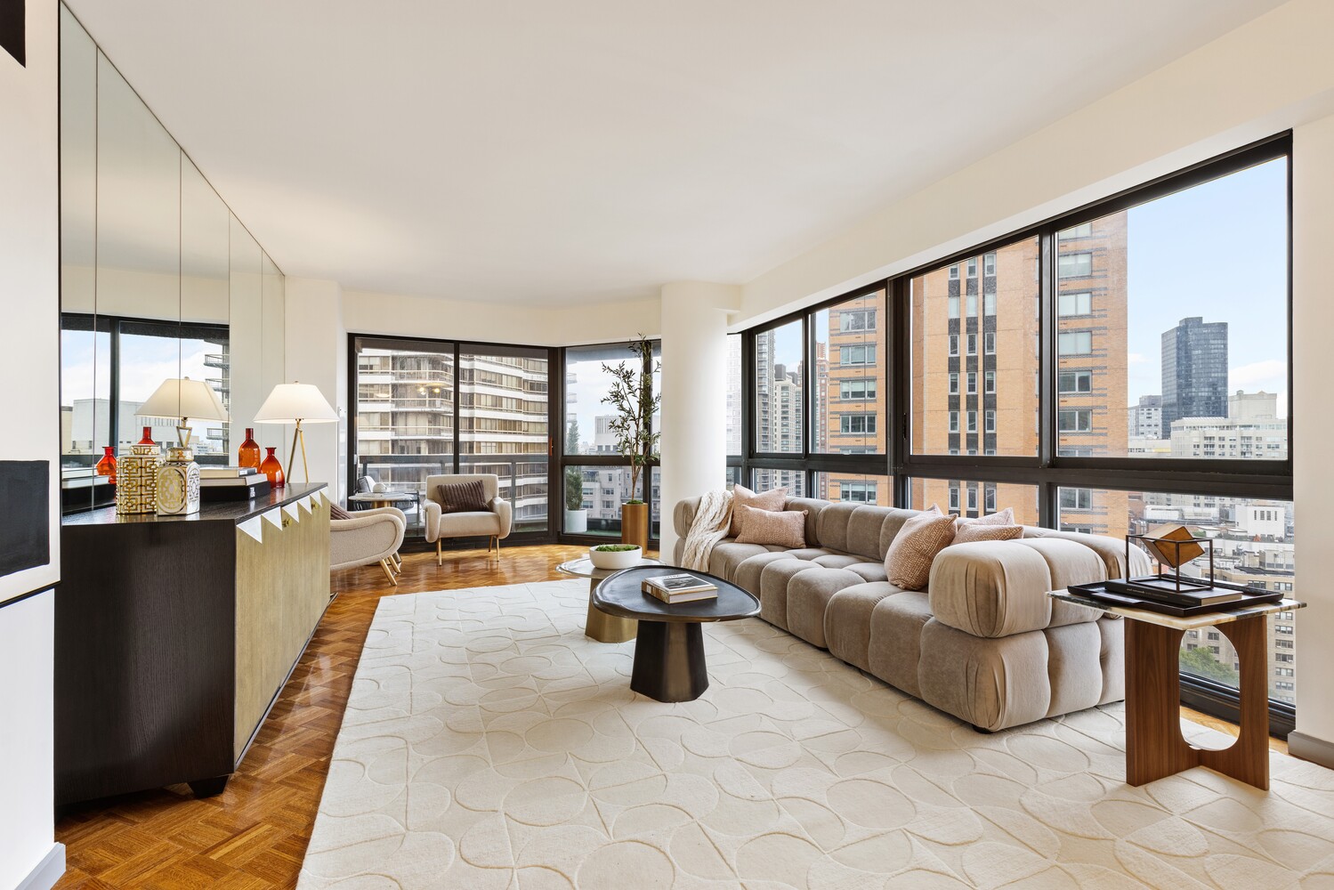 Furnished living room with a beige sectional, parquet floors, and broad windows overlooking city buildings.