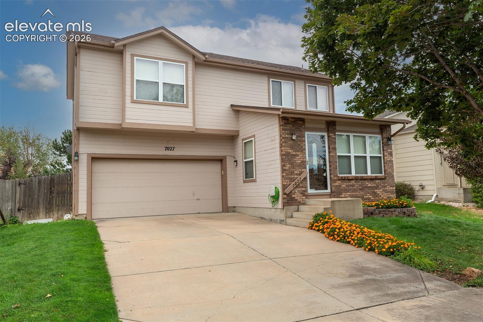 Two-story beige house with a double garage, brick entry, and orange flowers along the lawn.