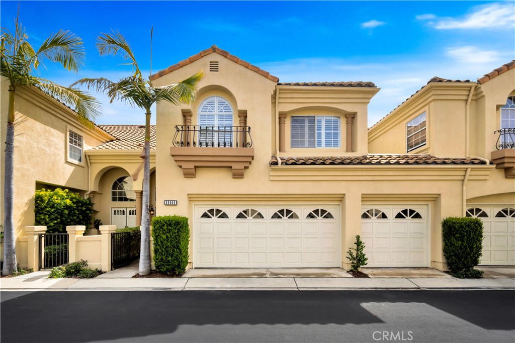 Two-story beige stucco home with tiled roof, balconies, palm trees, and two front-facing garage doors.