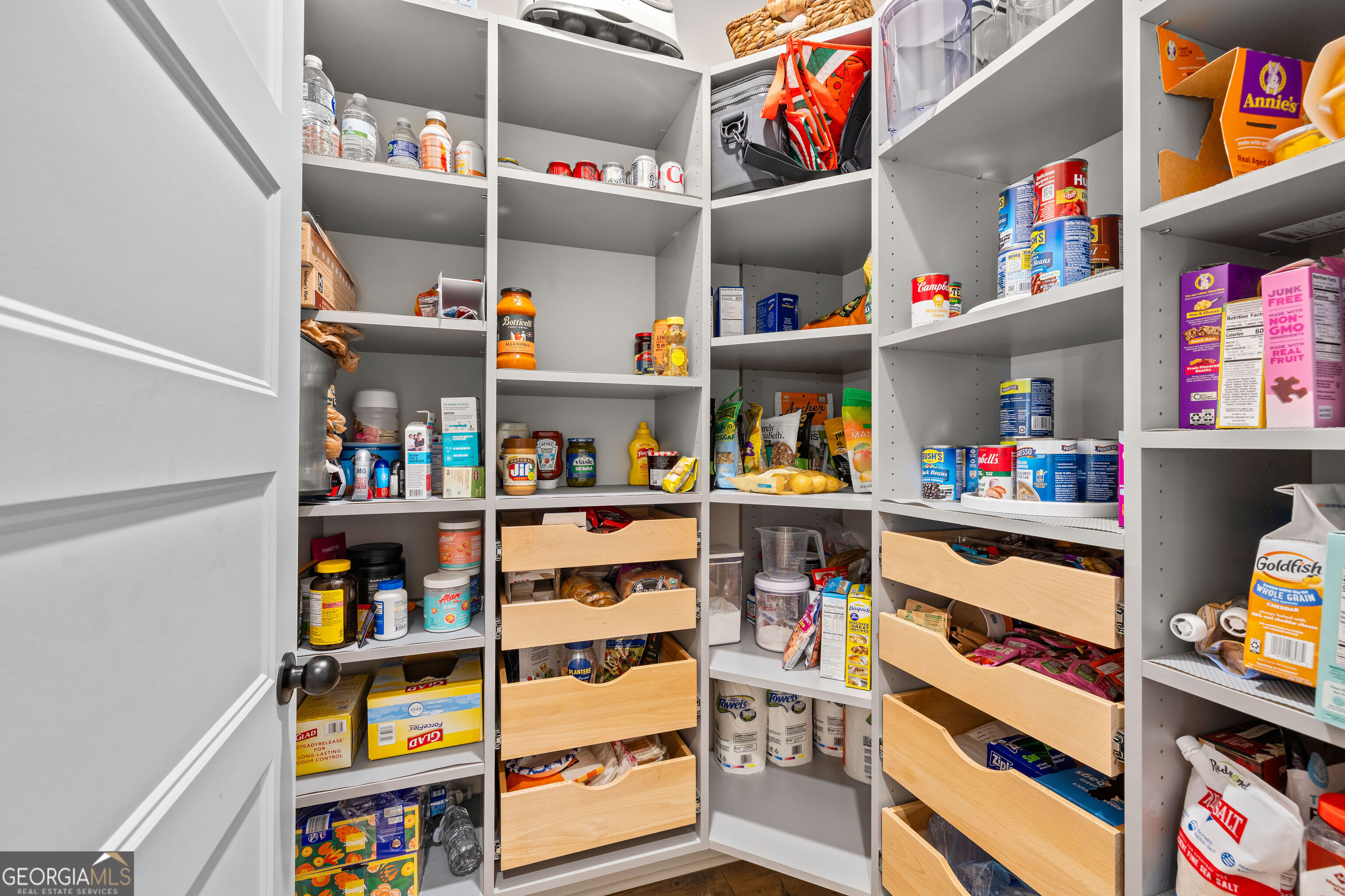 Walk-in pantry with stocked white shelves and light wood pull-out drawers.