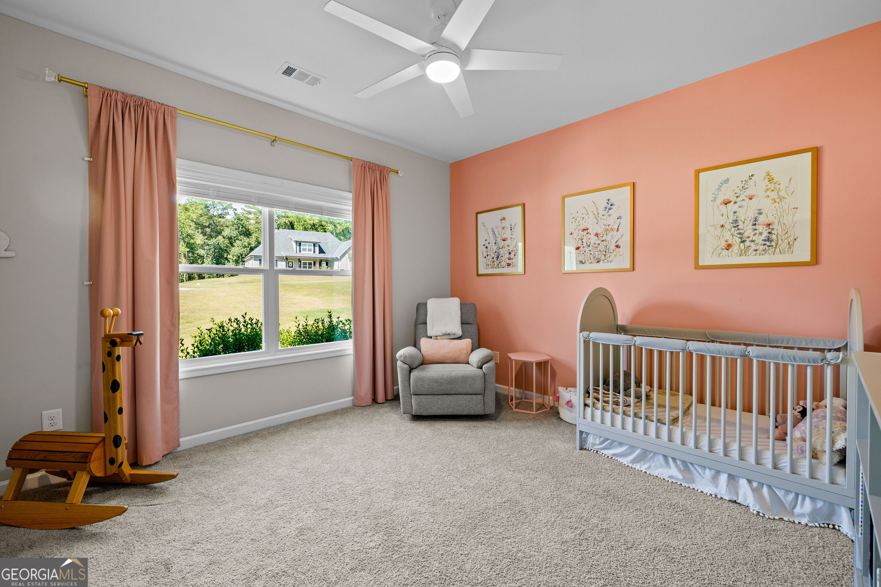 Nursery with a white crib, gray glider, coral walls, and a large window overlooking a lawn.