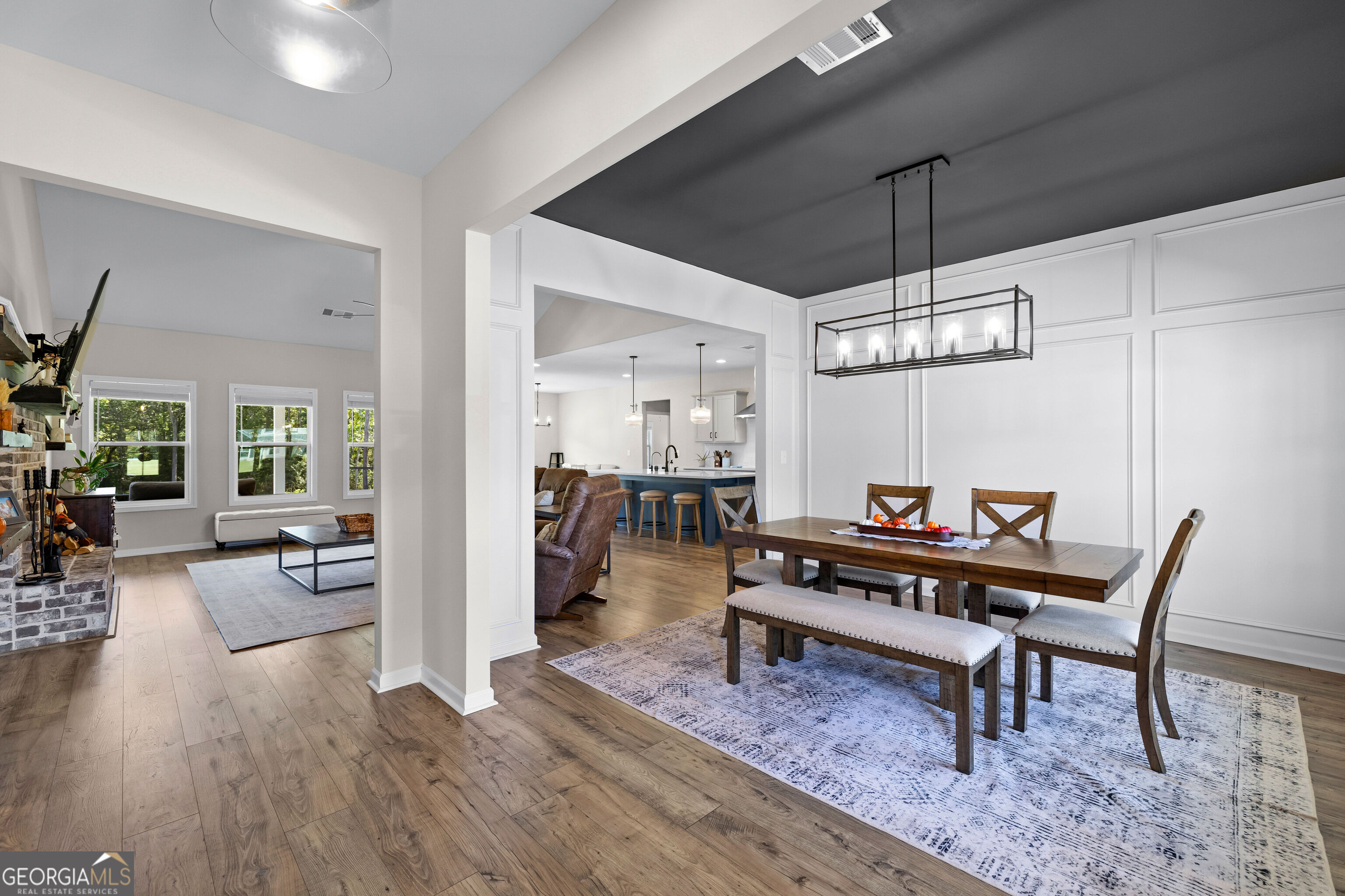 Open dining room with a large wood table, white paneled walls, and adjoining living room and kitchen.
