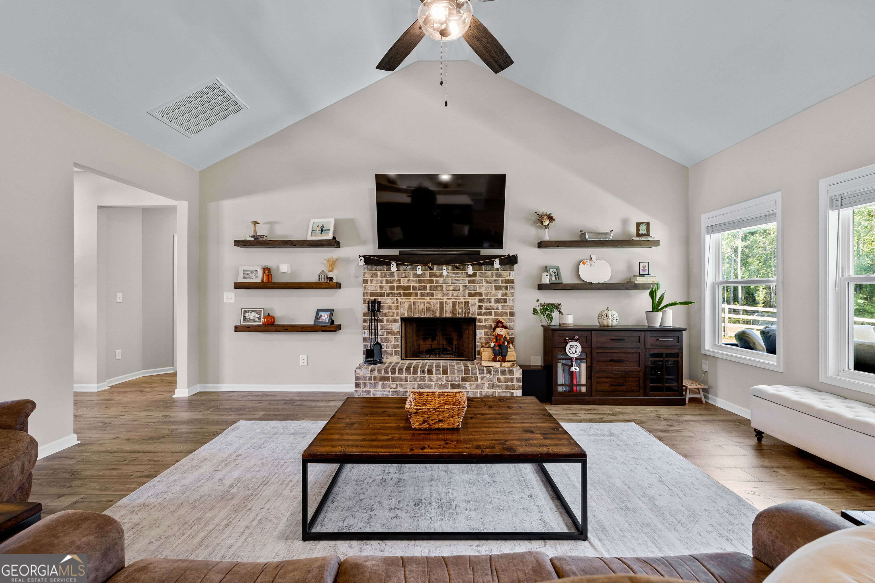 Living room with a vaulted ceiling, brick fireplace, mounted TV, and large windows overlooking trees.