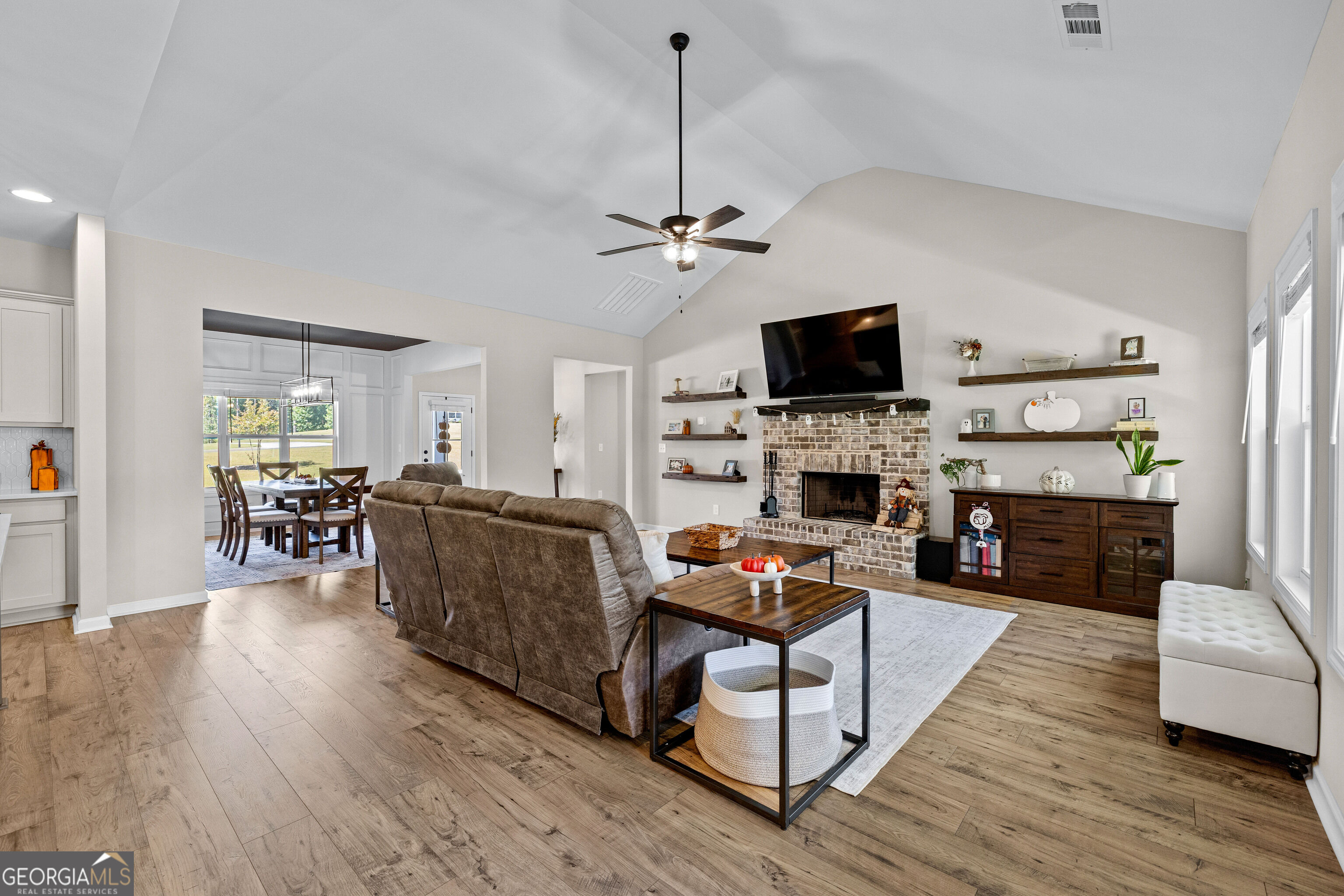 Open-plan living room with brick fireplace, mounted TV, wood floors, and adjoining dining area.
