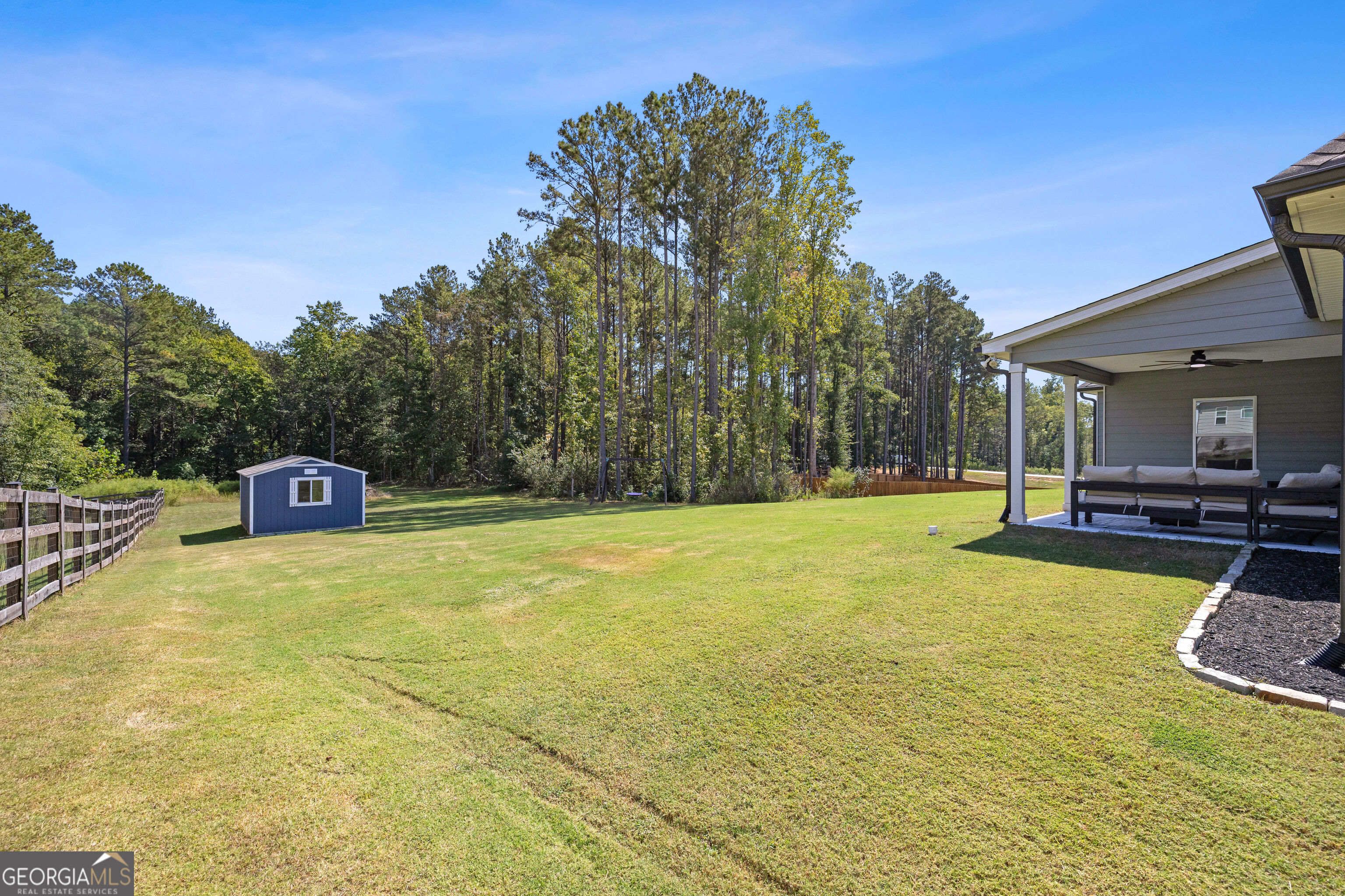 Spacious grassy backyard with a blue shed, wooden fence, wooded tree line, and covered patio.
