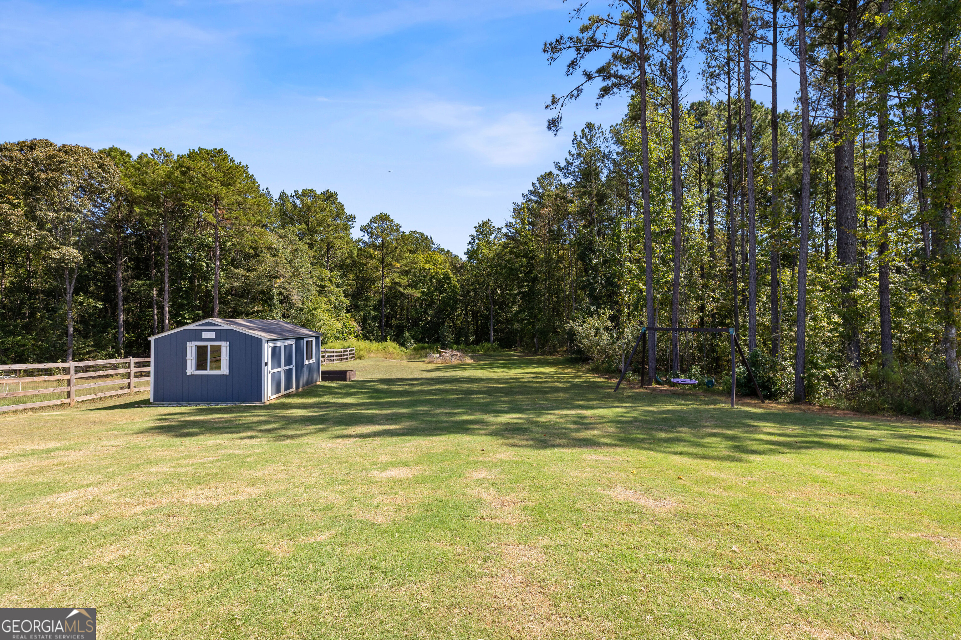 Grassy backyard with a blue storage shed, wooden fence, and swing set beneath mature trees.