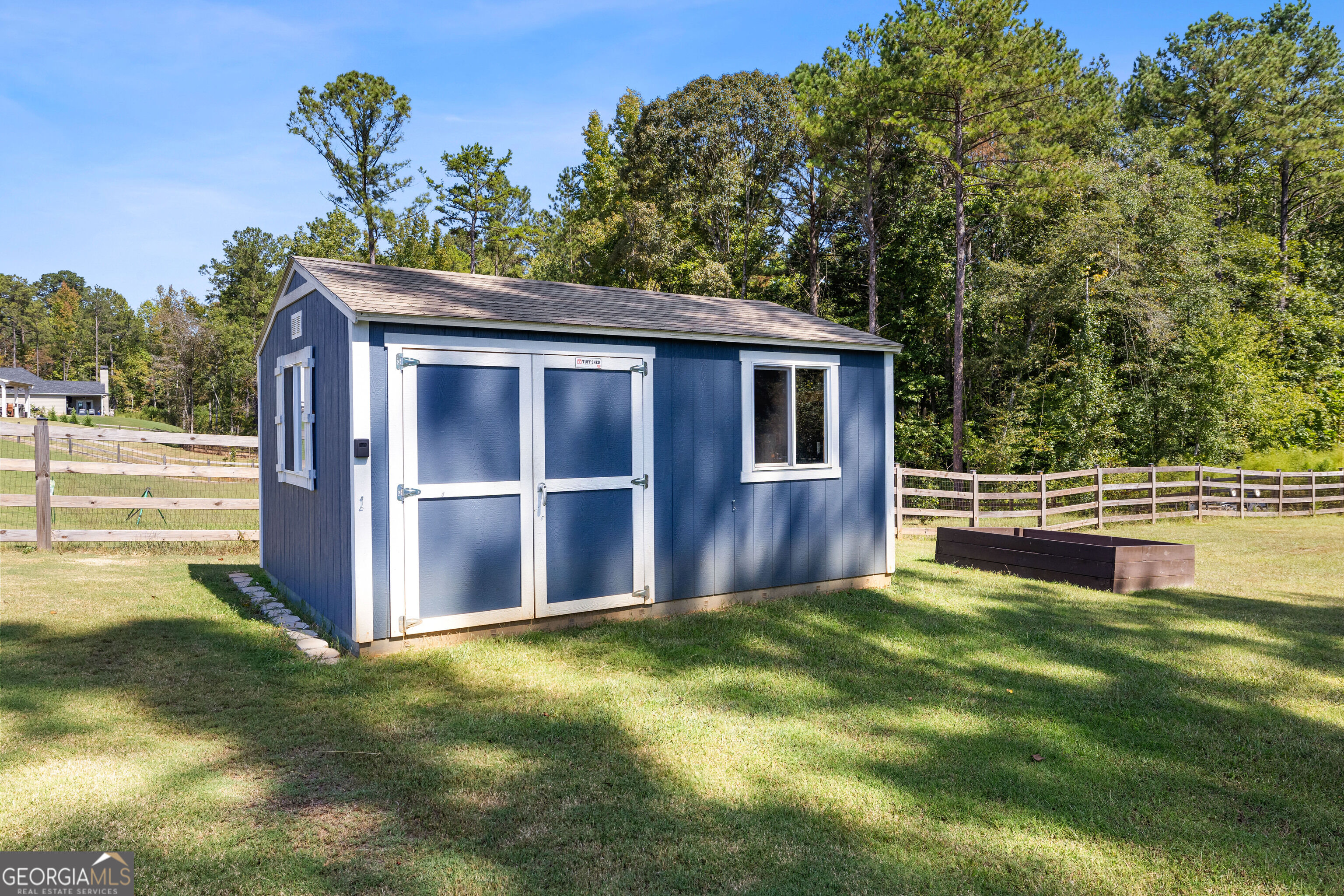 Blue storage shed with white trim in a fenced backyard beside a raised garden bed.