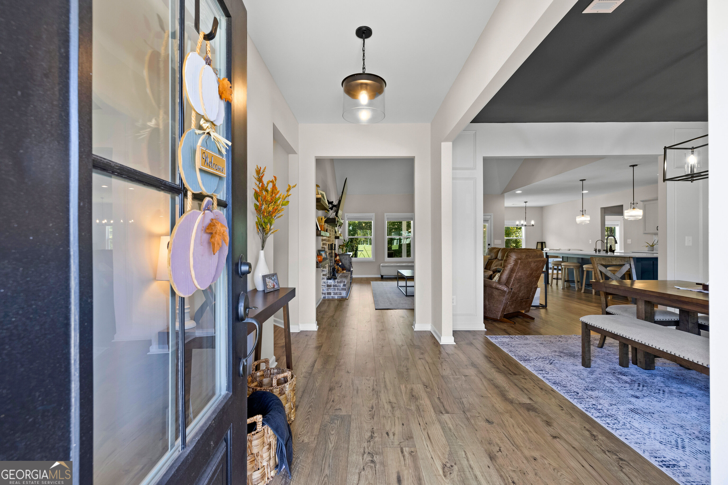 Open entryway with wood floors leading to furnished living, dining, and kitchen areas.