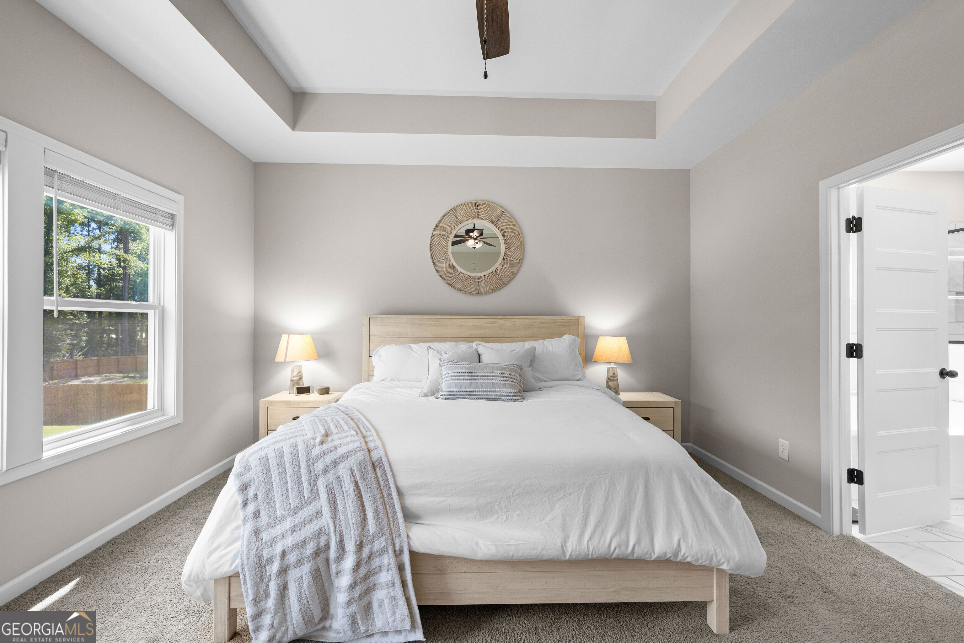 Furnished bedroom with a white bed, pale wood furniture, tray ceiling, and window overlooking trees.