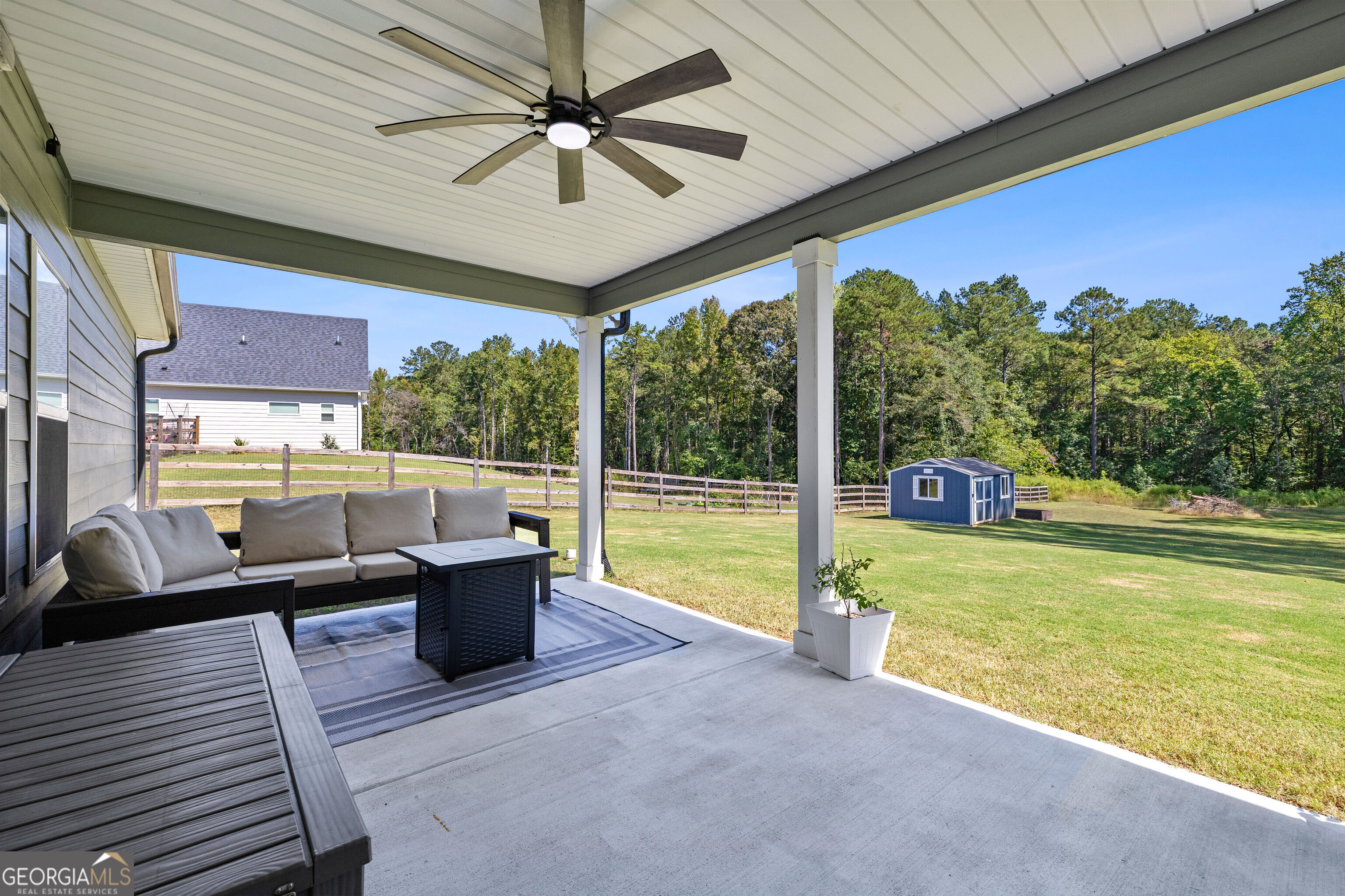Covered patio with outdoor seating and ceiling fan overlooking a fenced lawn and blue shed.