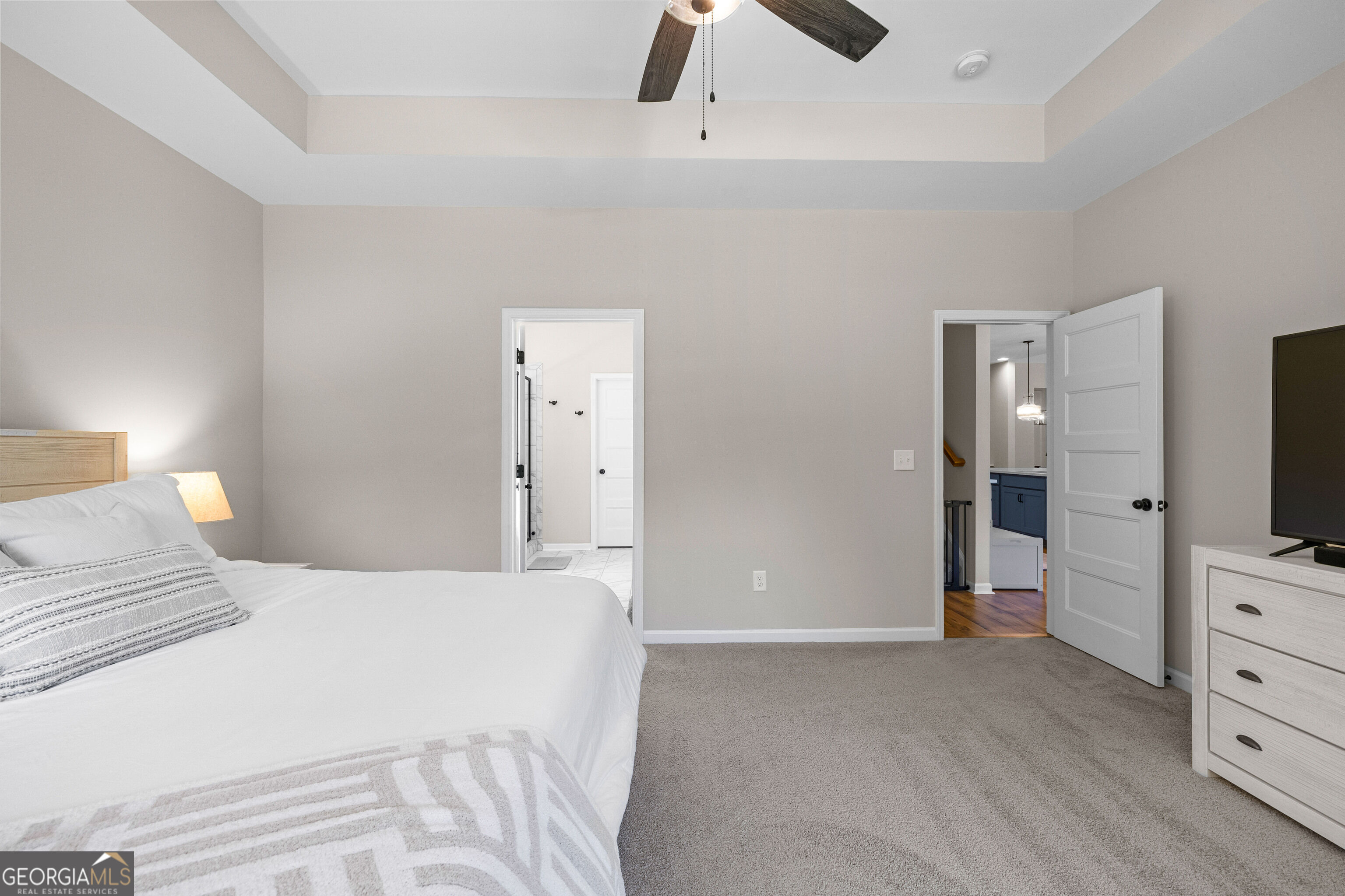 Furnished bedroom with a white bed, tray ceiling, and open doorways to a bathroom and hall.