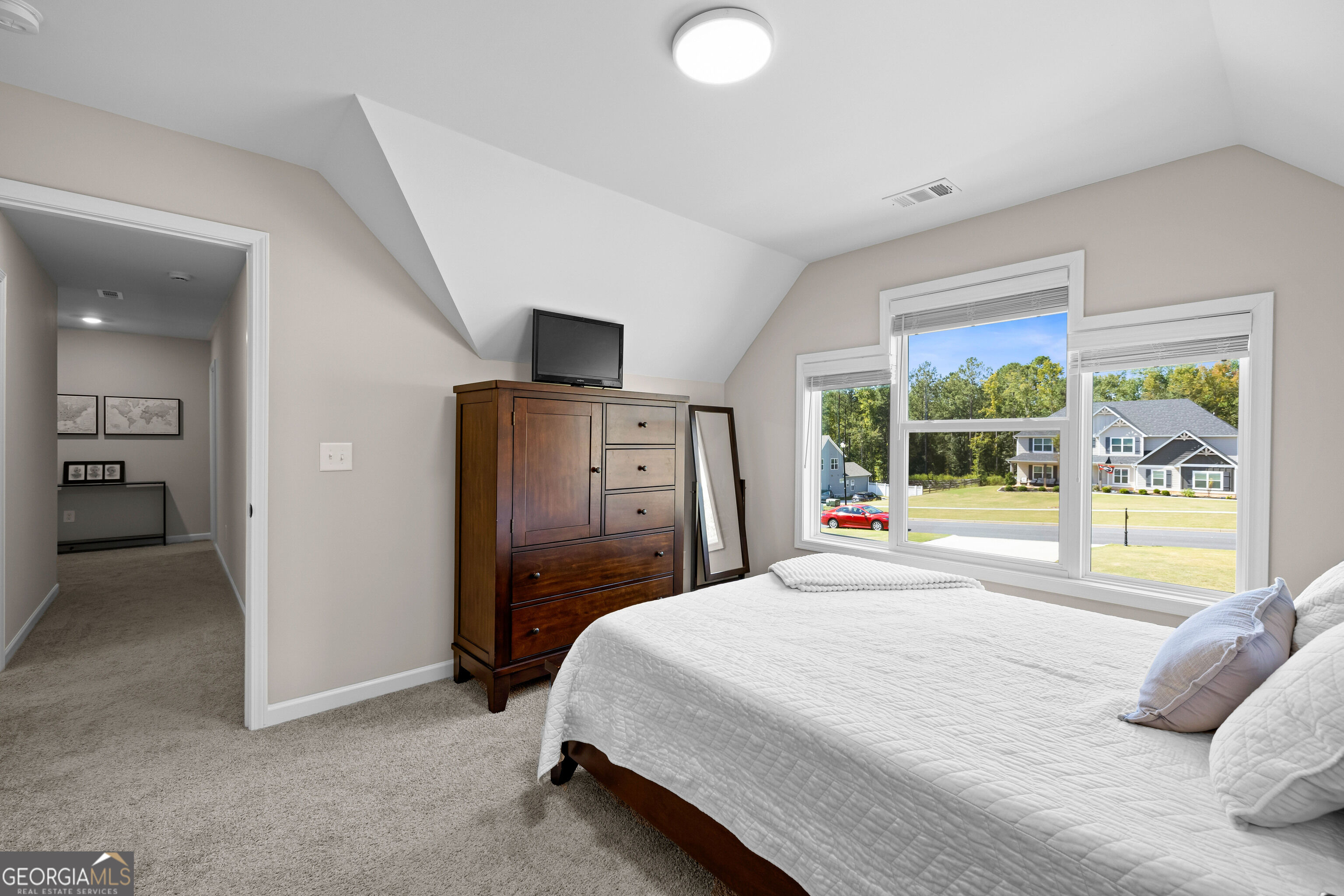Bedroom with a white quilted bed, dark wood dresser, sloped ceiling, and large windows overlooking neighboring homes.