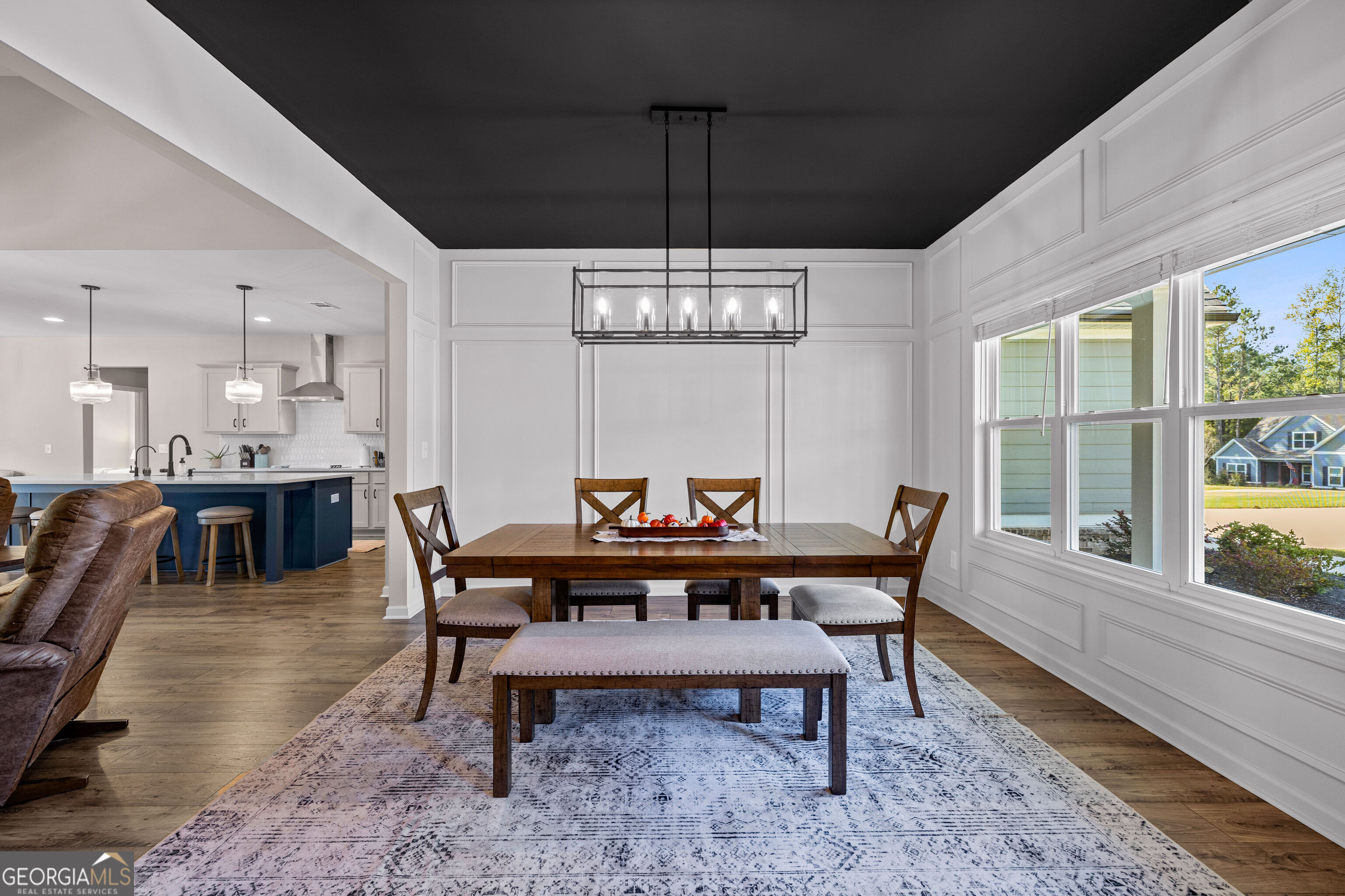 Dining room with a wooden table, rectangular chandelier, large windows, and an open kitchen.