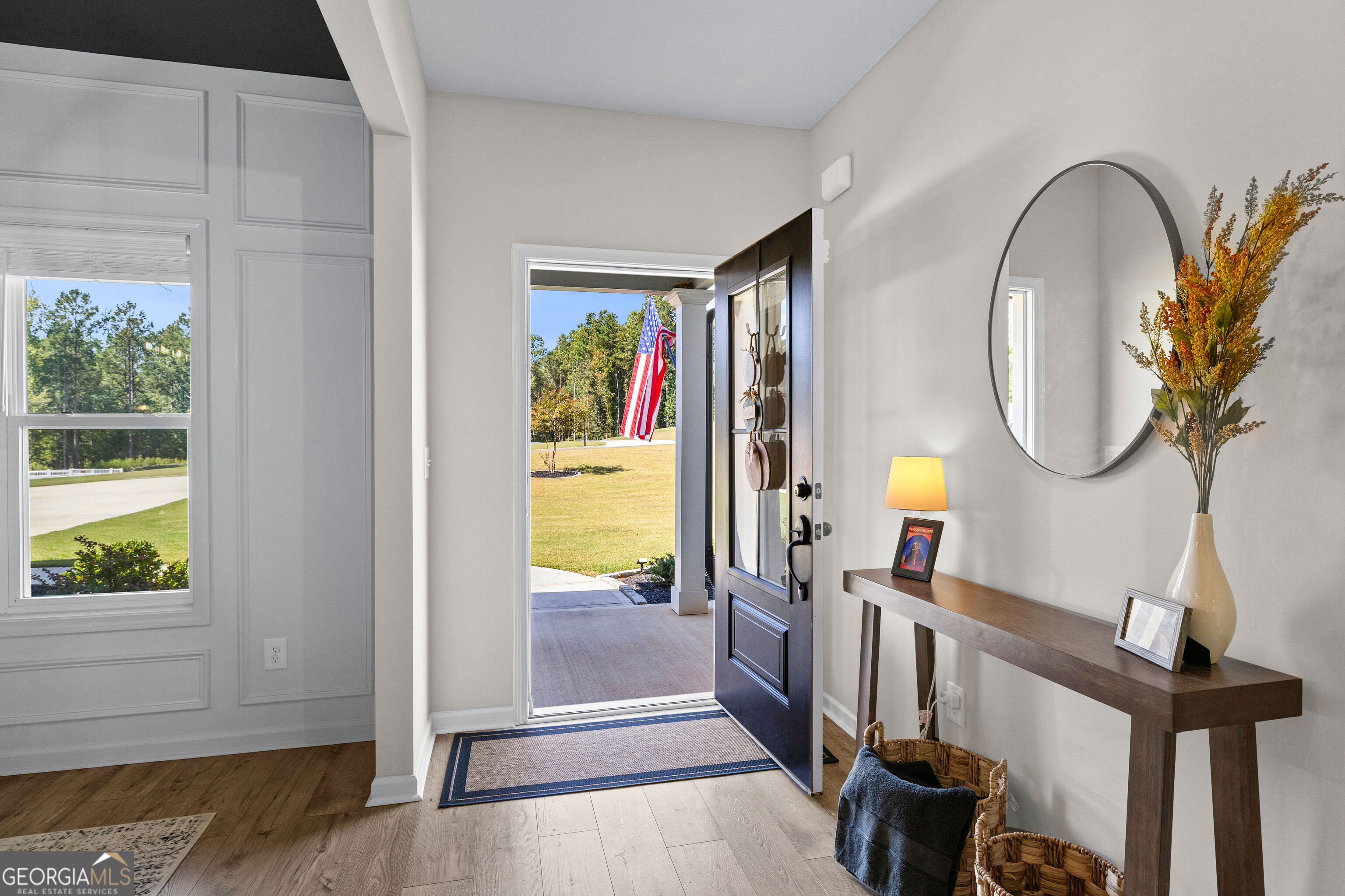Staged entryway with an open dark front door, paneled walls, and a console table beneath a round mirror.