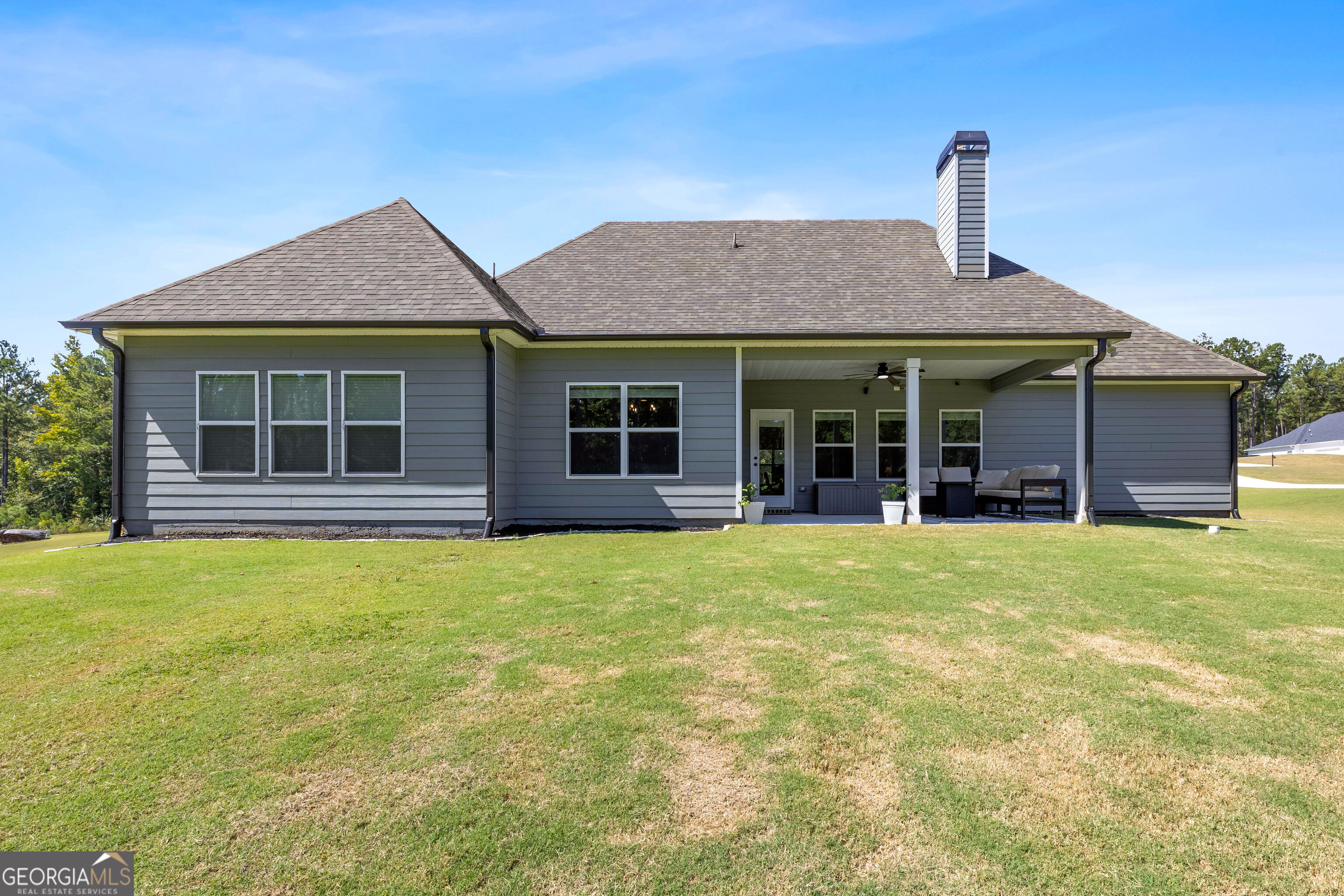 Rear of a gray house with a covered patio, outdoor seating, and a broad lawn.