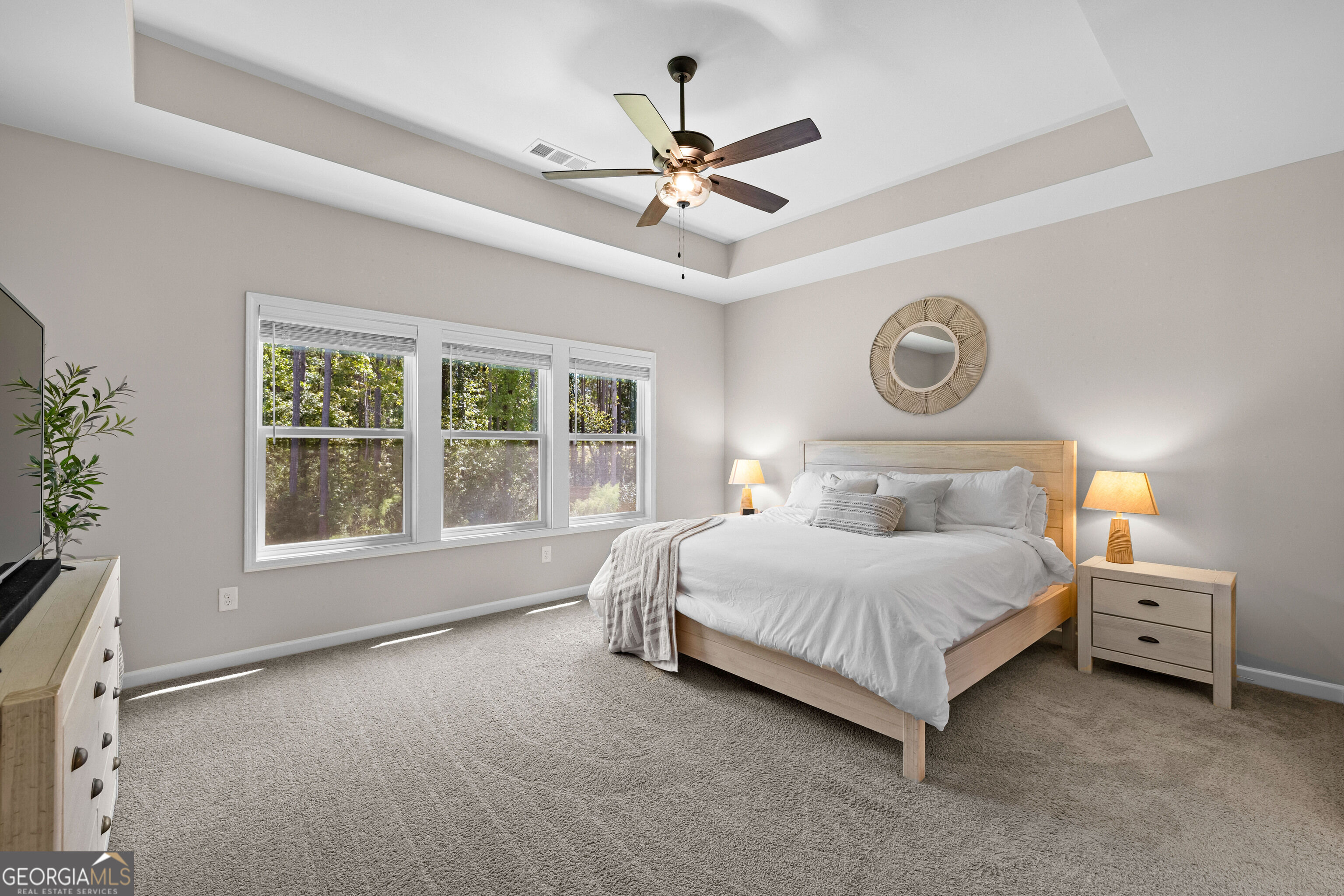 Staged bedroom with a white bed, tray ceiling, ceiling fan, and three windows overlooking trees.