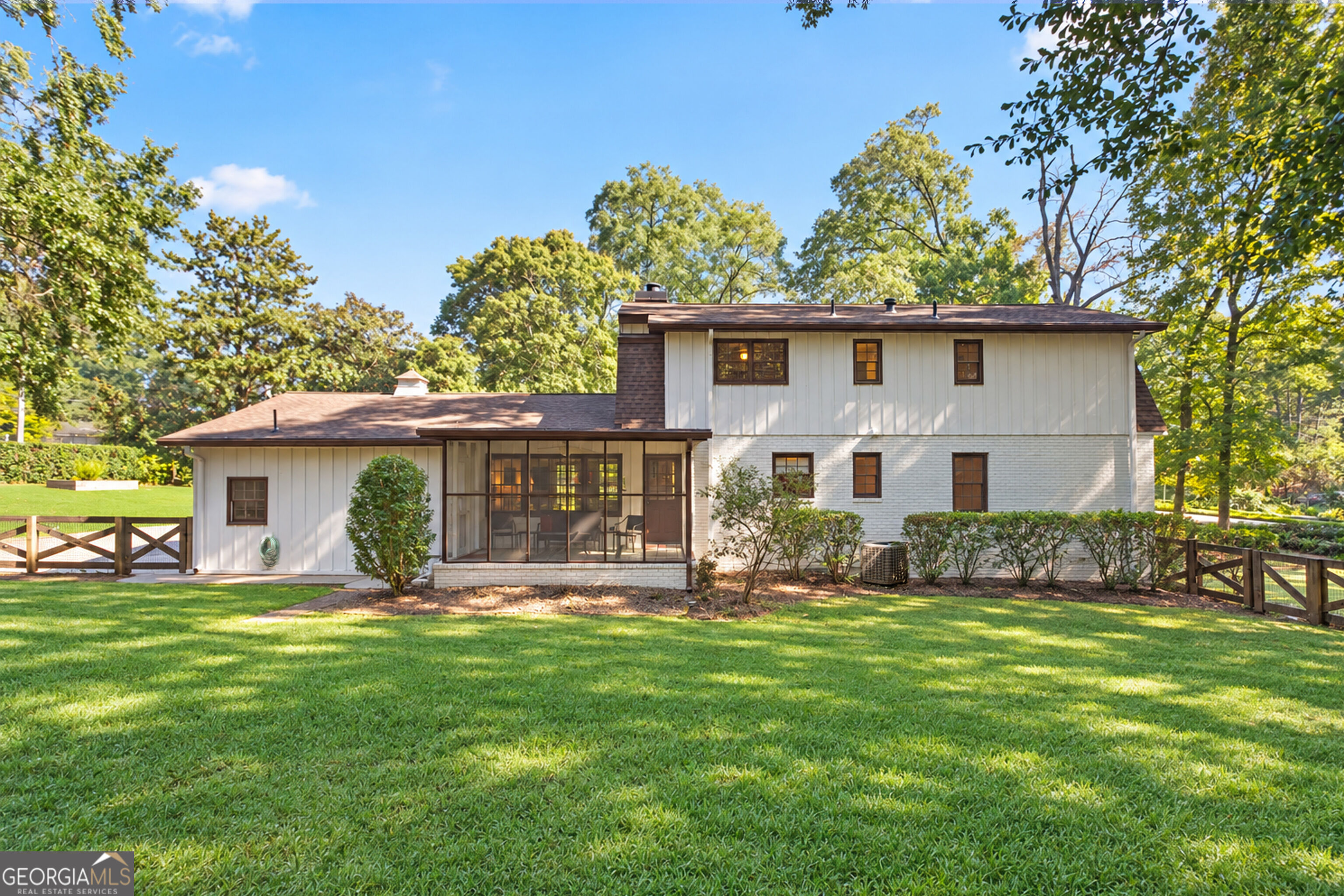 Two-story white-and-brick house with a screened porch, broad lawn, mature trees, and wooden fencing.