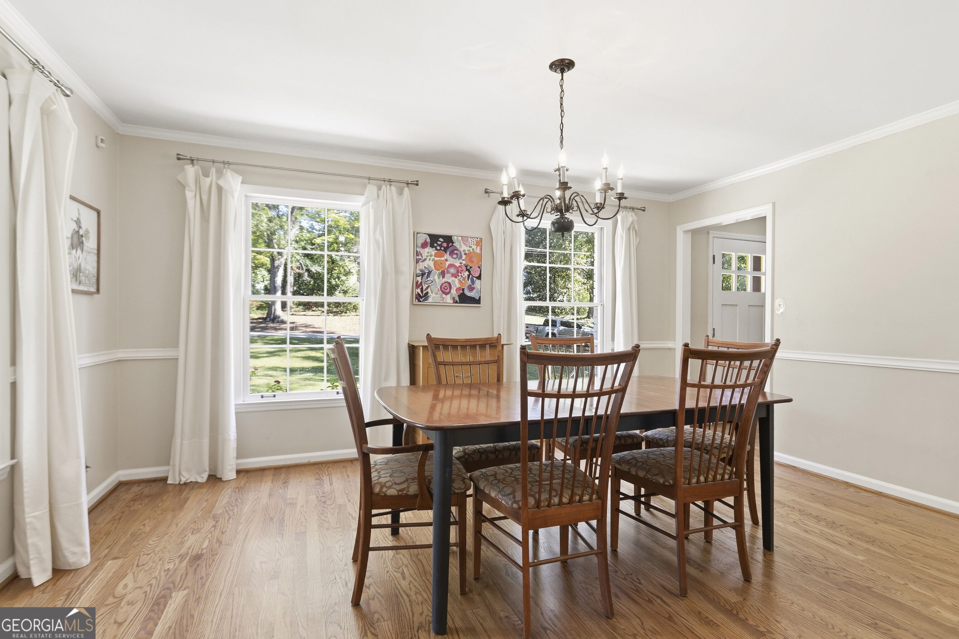 Dining room with a wooden table, hardwood floors, large curtained windows, and a chandelier.