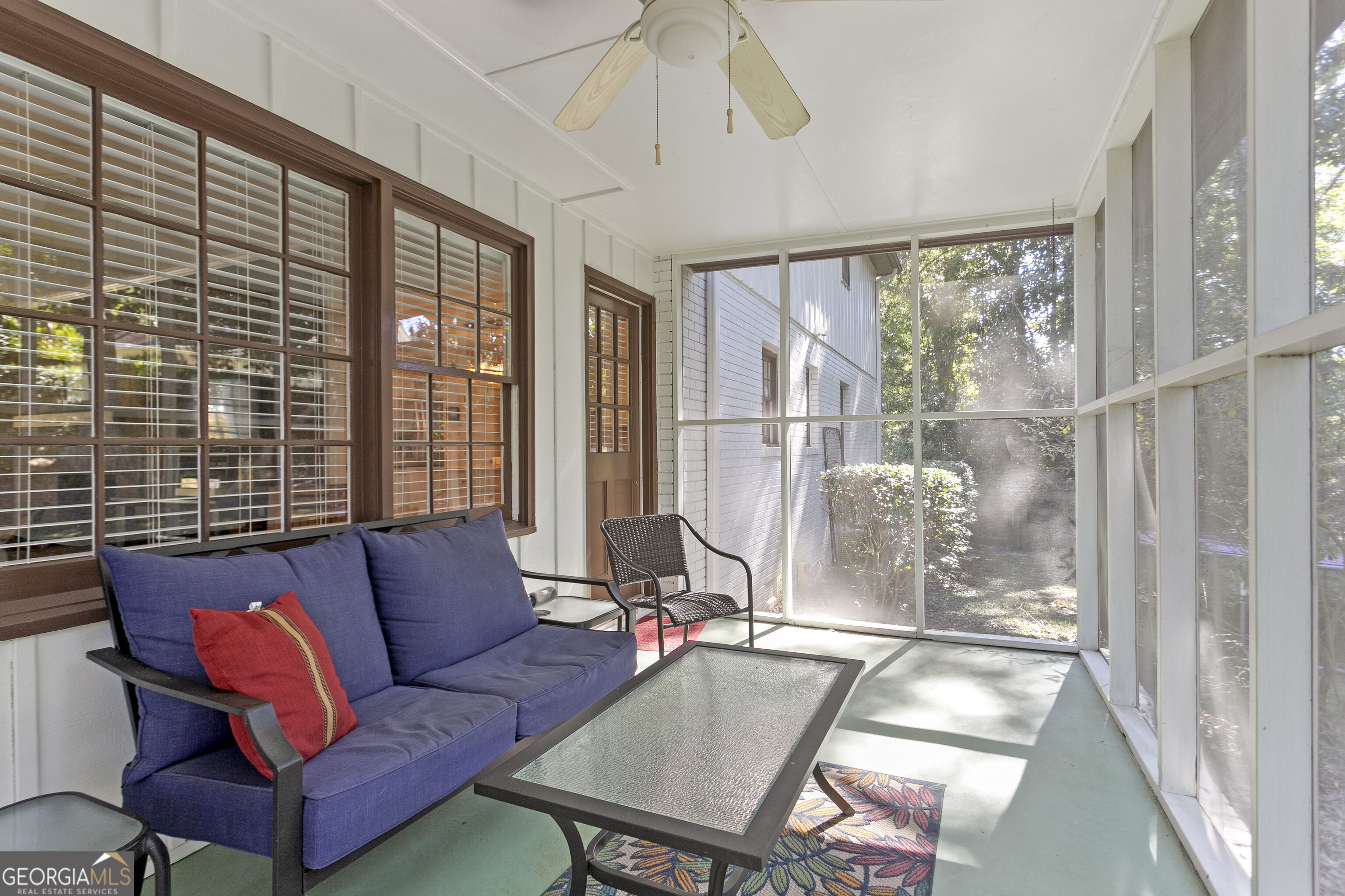 Screened porch with blue sofa, red pillow, glass-top table, and leafy views.