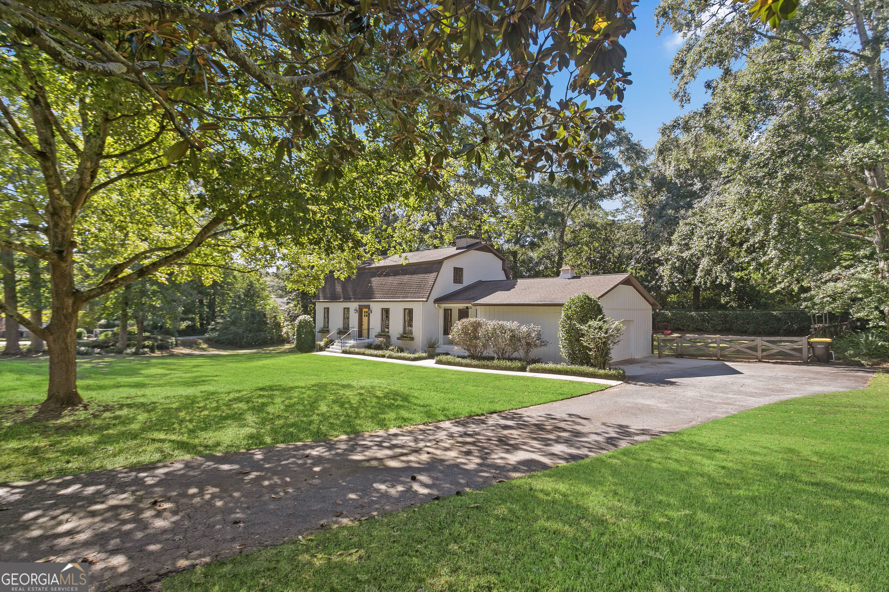 Light-colored house with an attached garage, broad lawn, mature shade trees, and paved driveway.