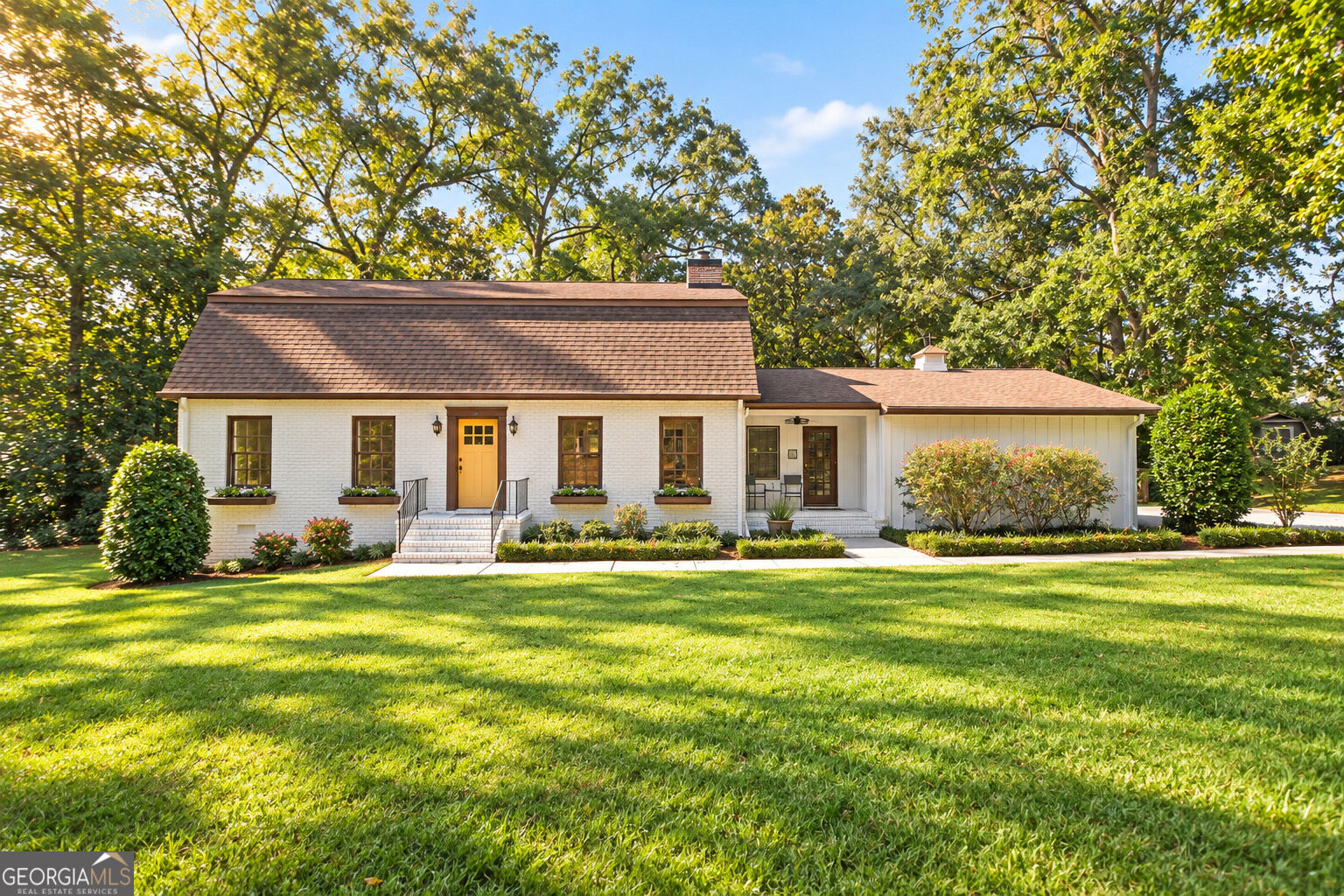 White brick home with a yellow front door, landscaped shrubs, broad lawn, and mature trees.