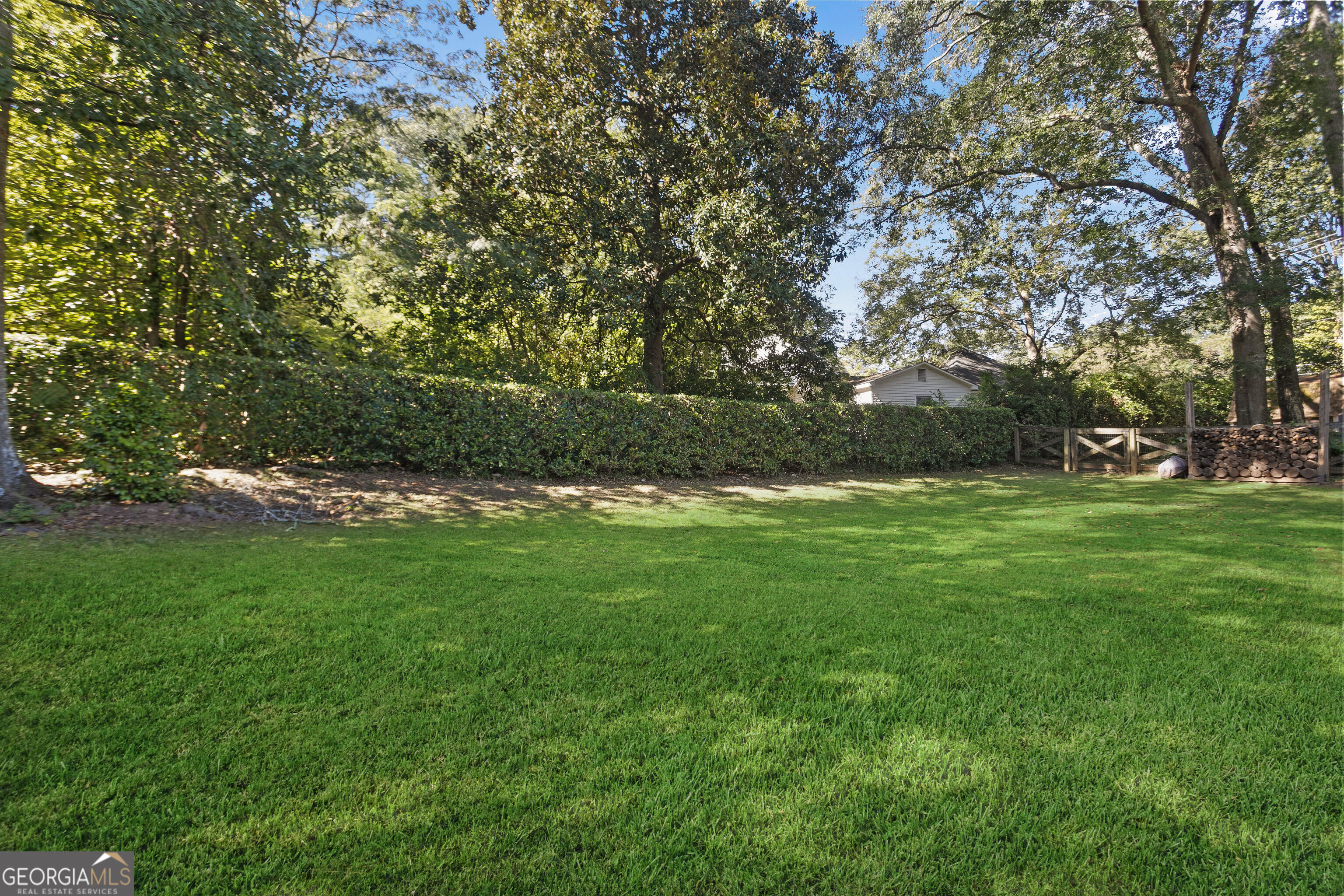 Backyard with a broad lawn, mature shade trees, tall hedge, and wooden gate beside stacked firewood.