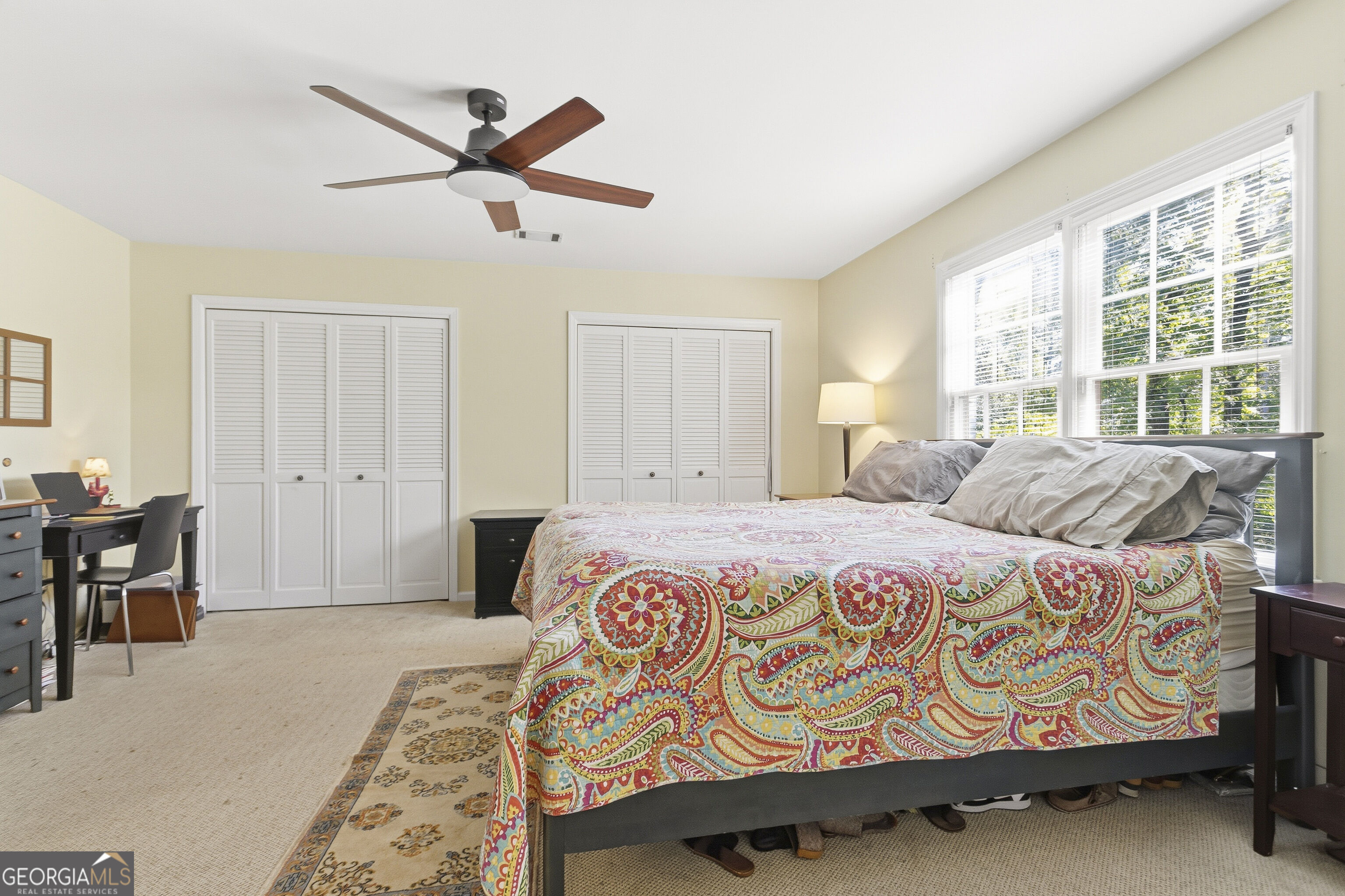 Bedroom with a colorful patterned quilt, white closet doors, and large sunlit windows.