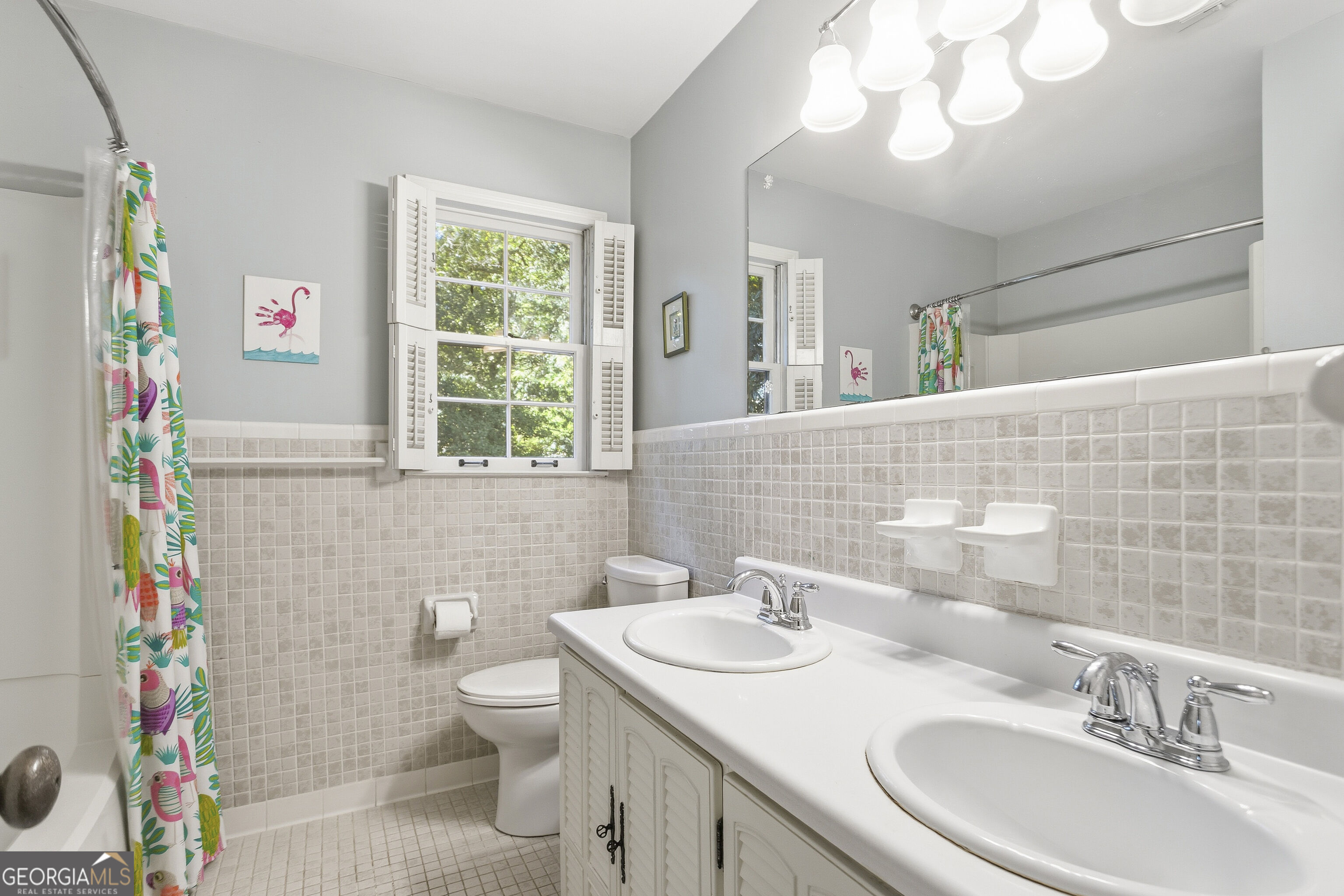 Bathroom with double vanity, beige tile, shuttered window, and colorful shower curtain.