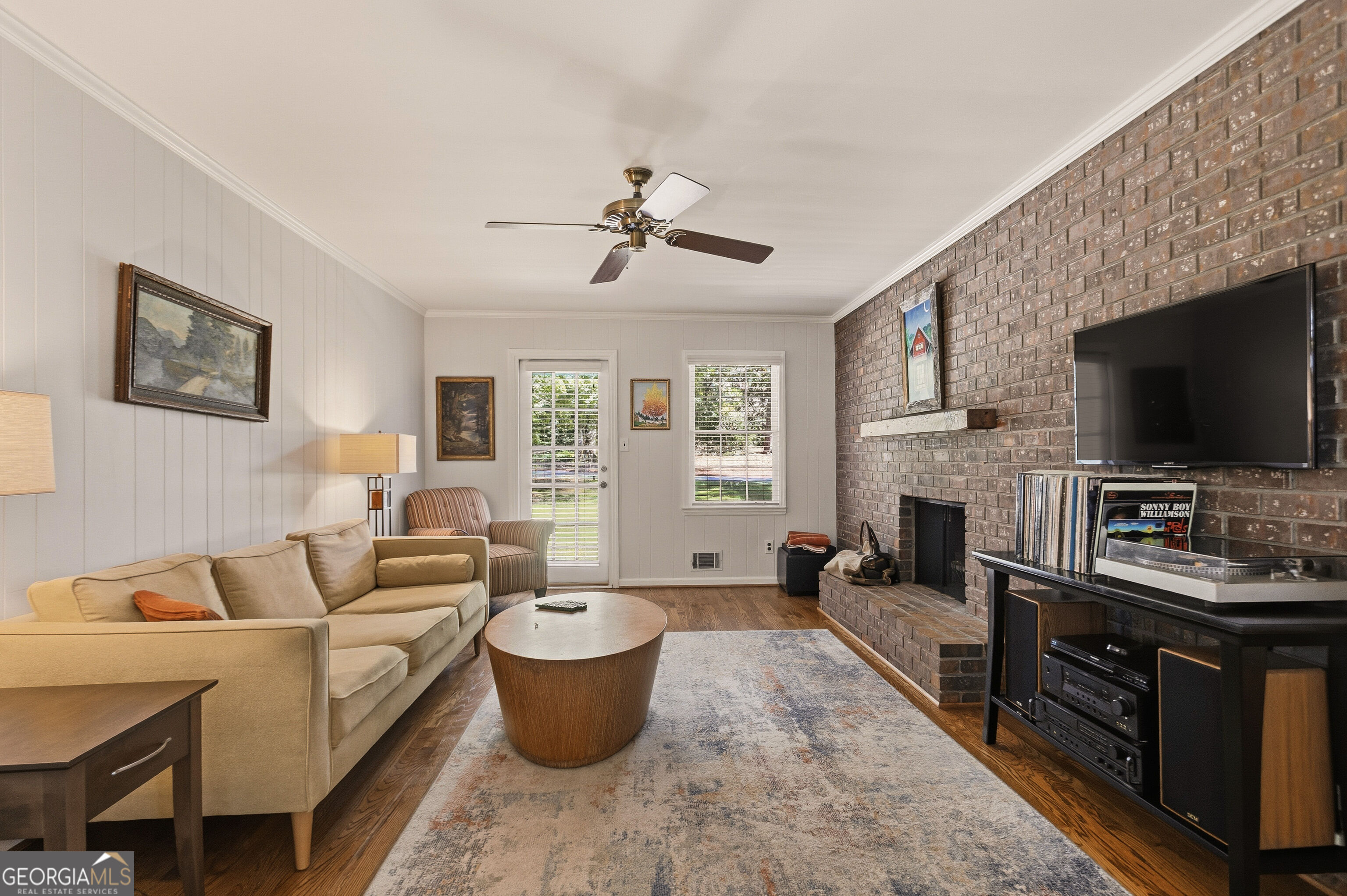 Living room with a brick fireplace wall, beige seating, television, and hardwood floors.
