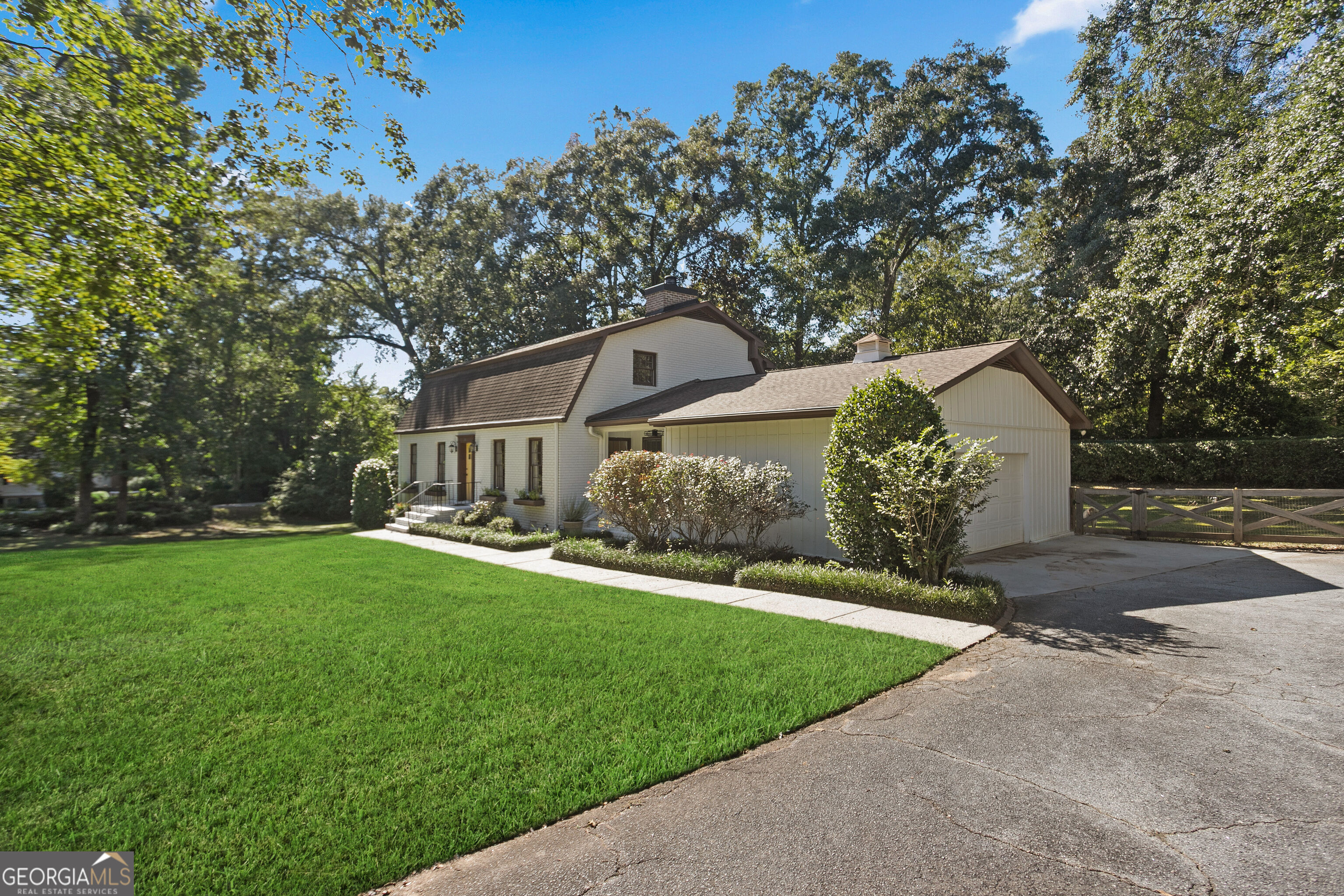 Light-colored house with an attached garage, broad lawn, mature trees, and paved driveway.