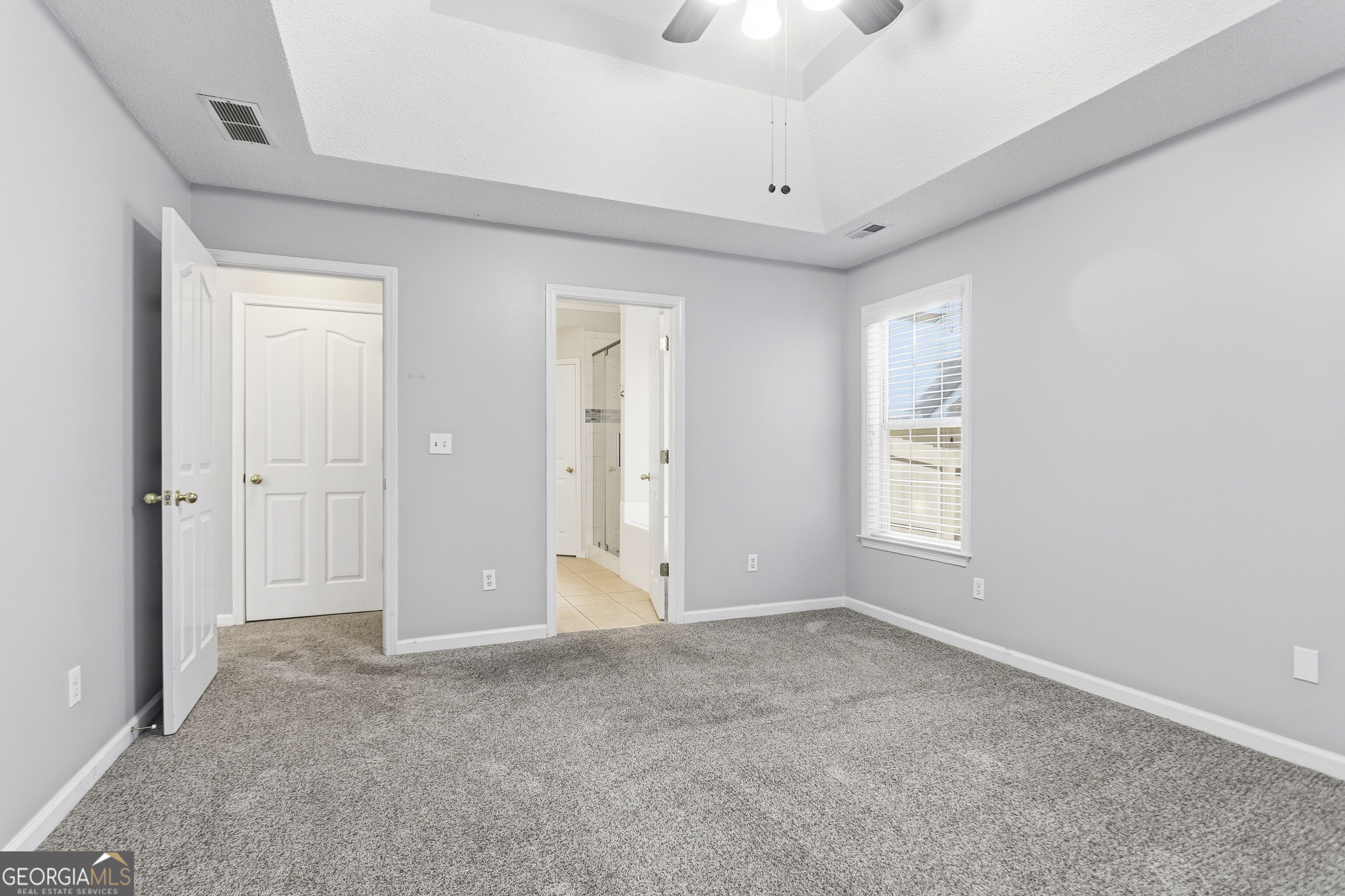 Empty bedroom with gray carpet, a tray ceiling, and a doorway to an ensuite bathroom.