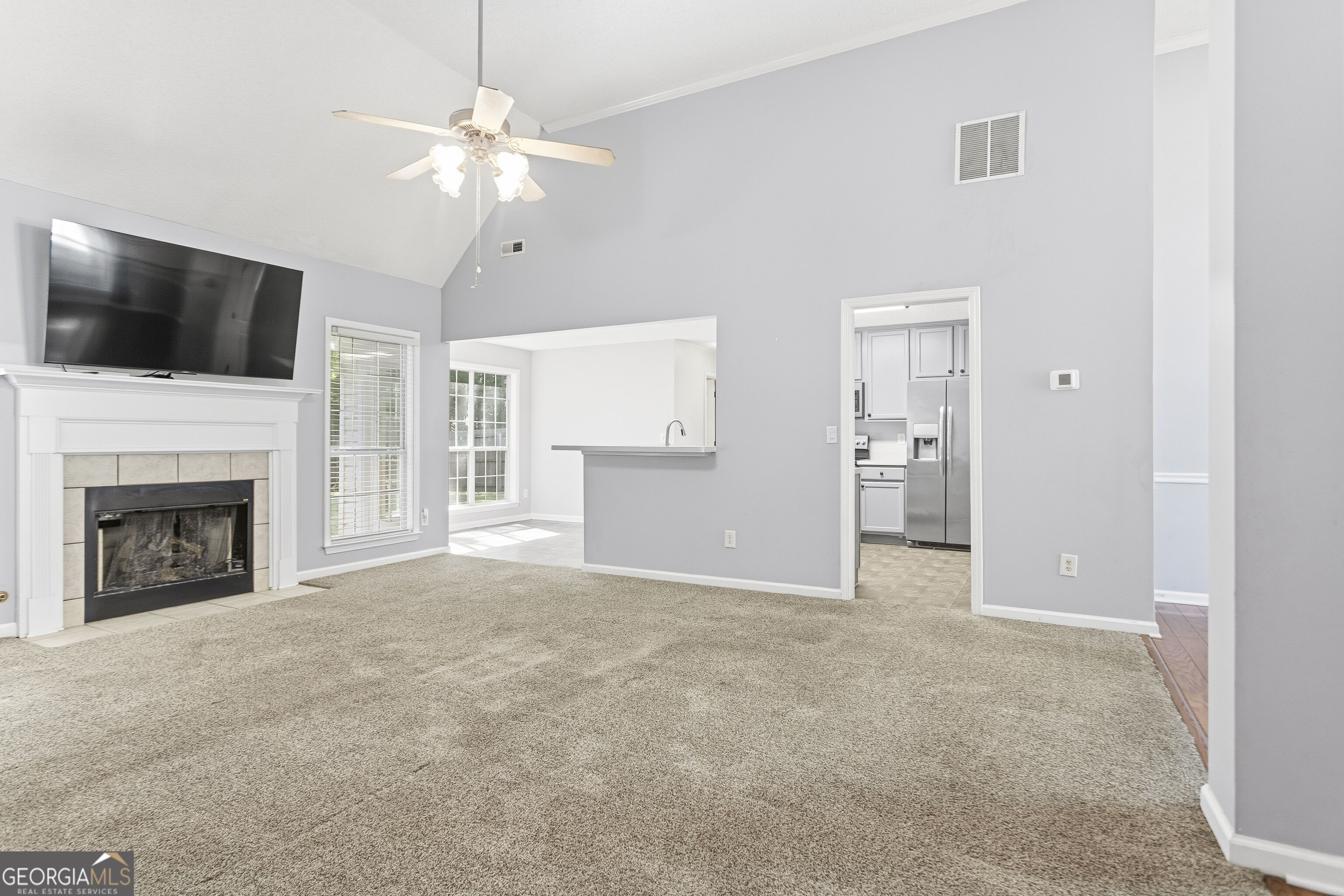 Living room with vaulted ceilings, beige carpet, a white fireplace, and an open view toward the kitchen.