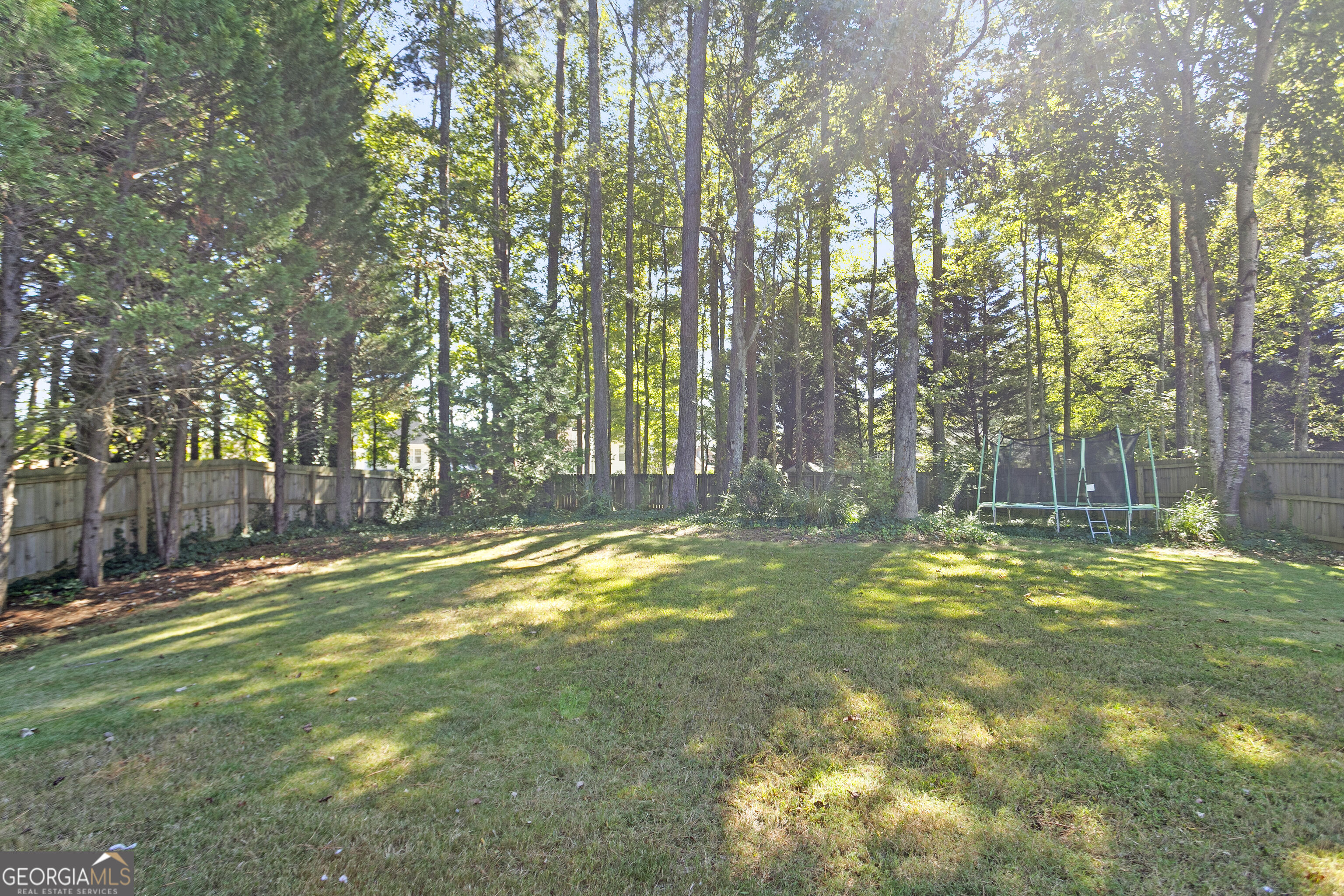 Fenced grassy backyard shaded by tall trees, with a trampoline near the back fence.
