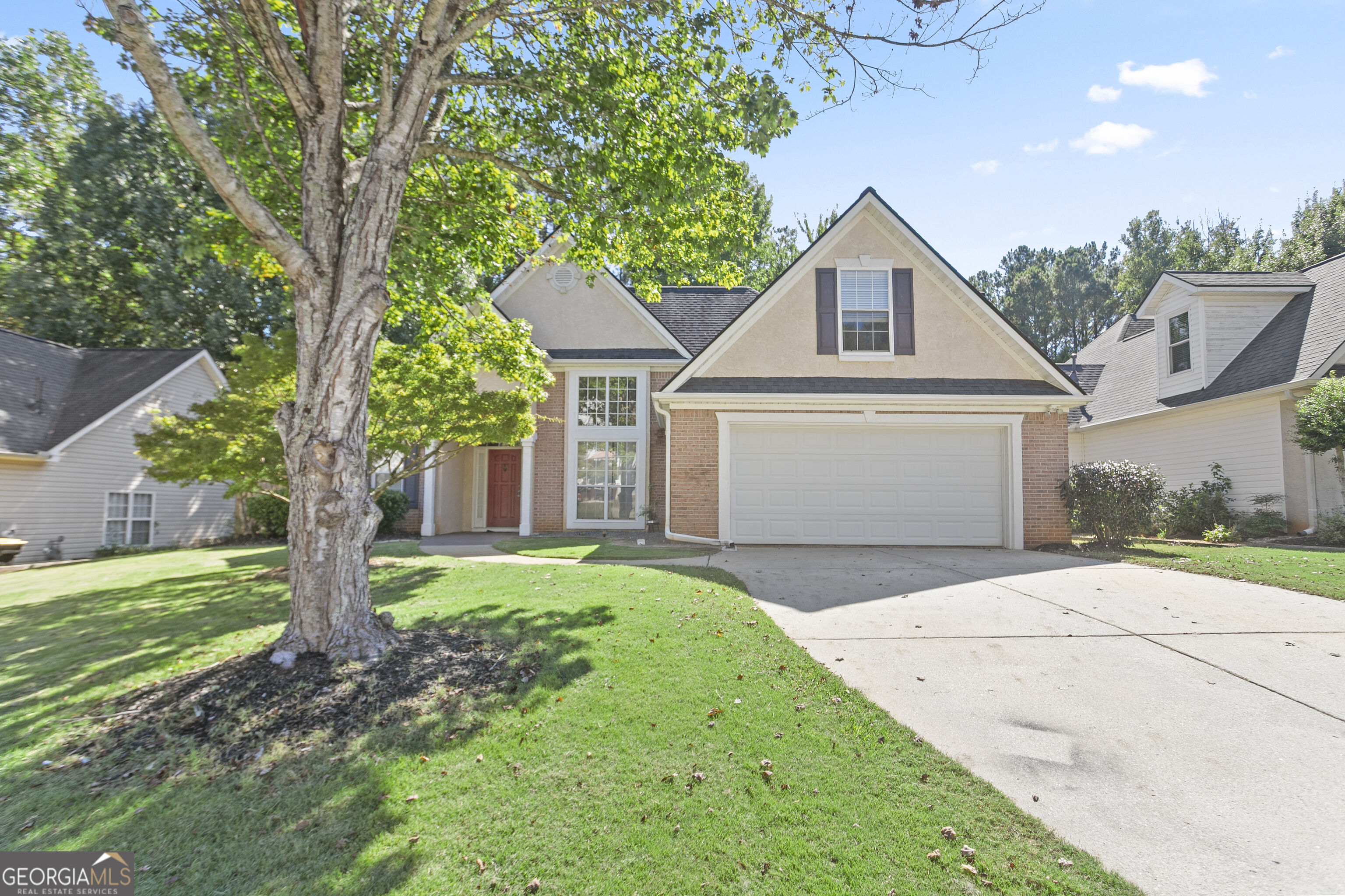 Two-story brick-and-stucco house with a double garage, broad driveway, and large shade tree in front.
