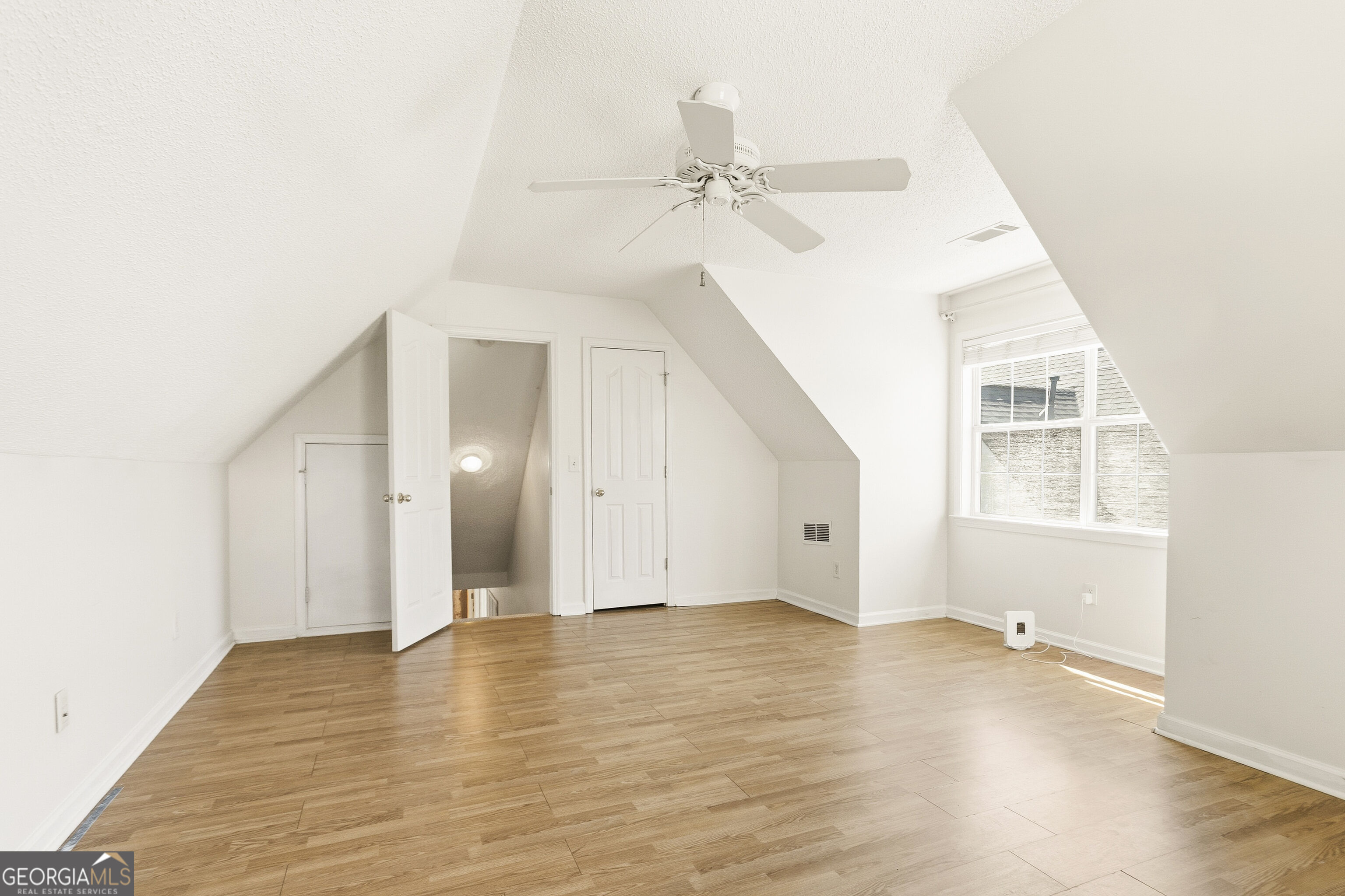 Empty upper-floor room with sloped ceilings, wood flooring, a ceiling fan, and a bright window.