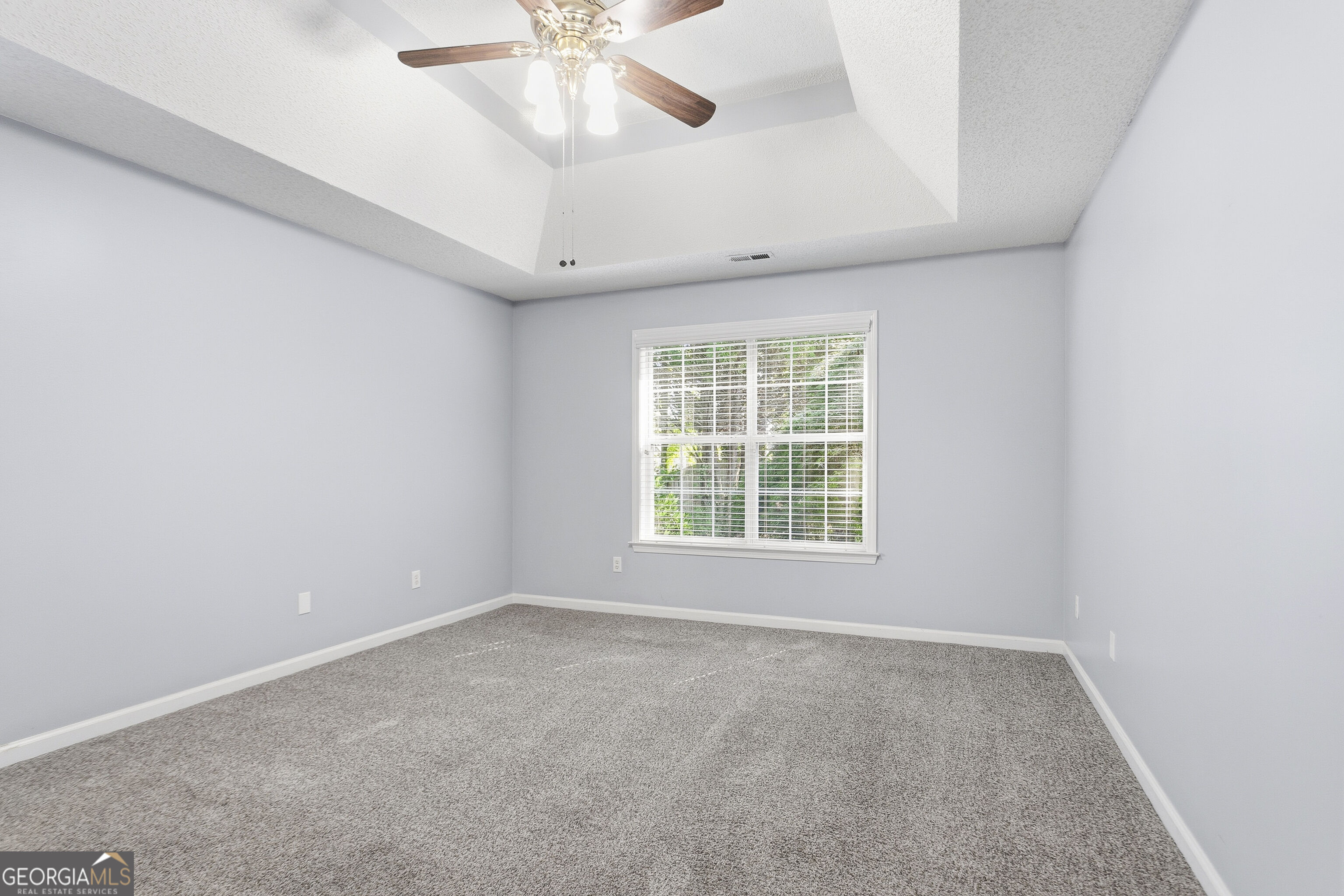 Empty carpeted bedroom with a tray ceiling, ceiling fan, and window with white blinds.