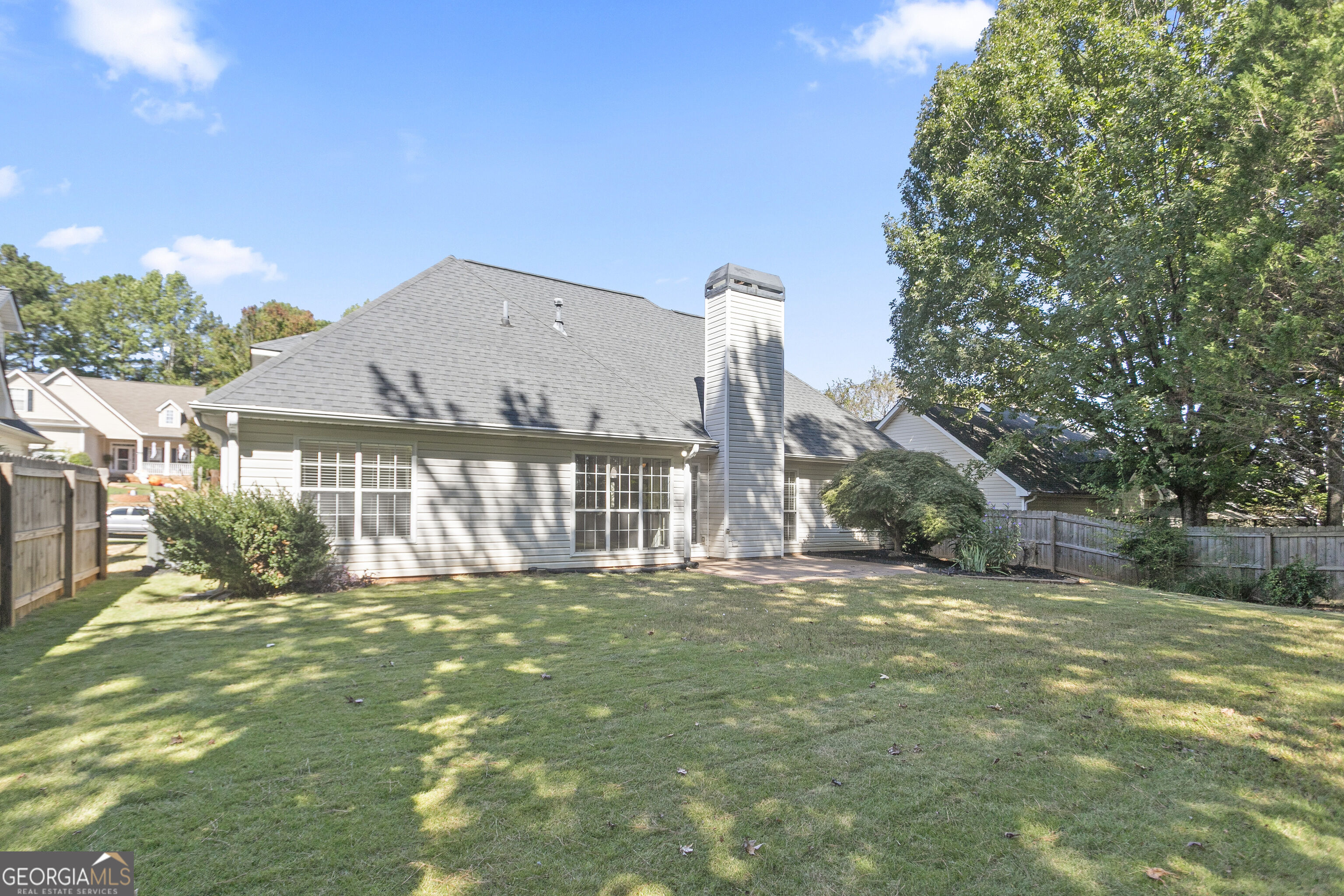 Rear of a gray-sided house with a tall chimney, small patio, and fenced lawn beneath mature trees.