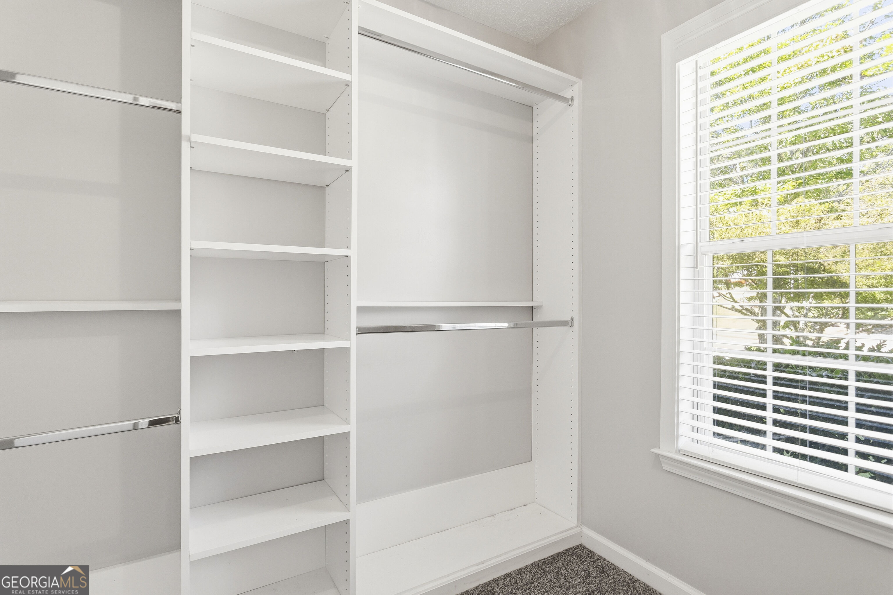 Walk-in closet with white built-in shelves, hanging rods, and a large window with blinds.