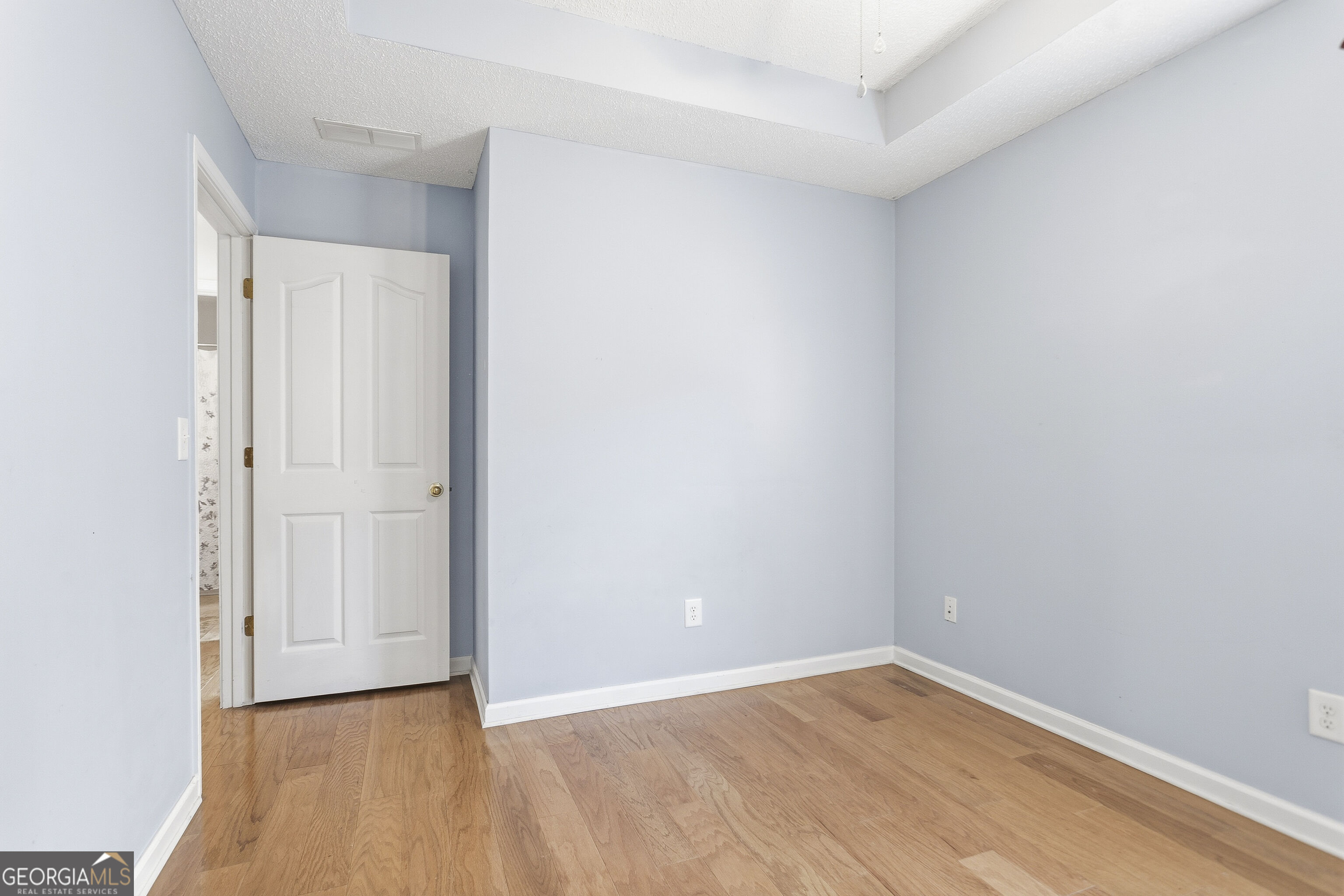 Empty room with pale blue walls, hardwood floors, and an open white door beneath a stepped ceiling.