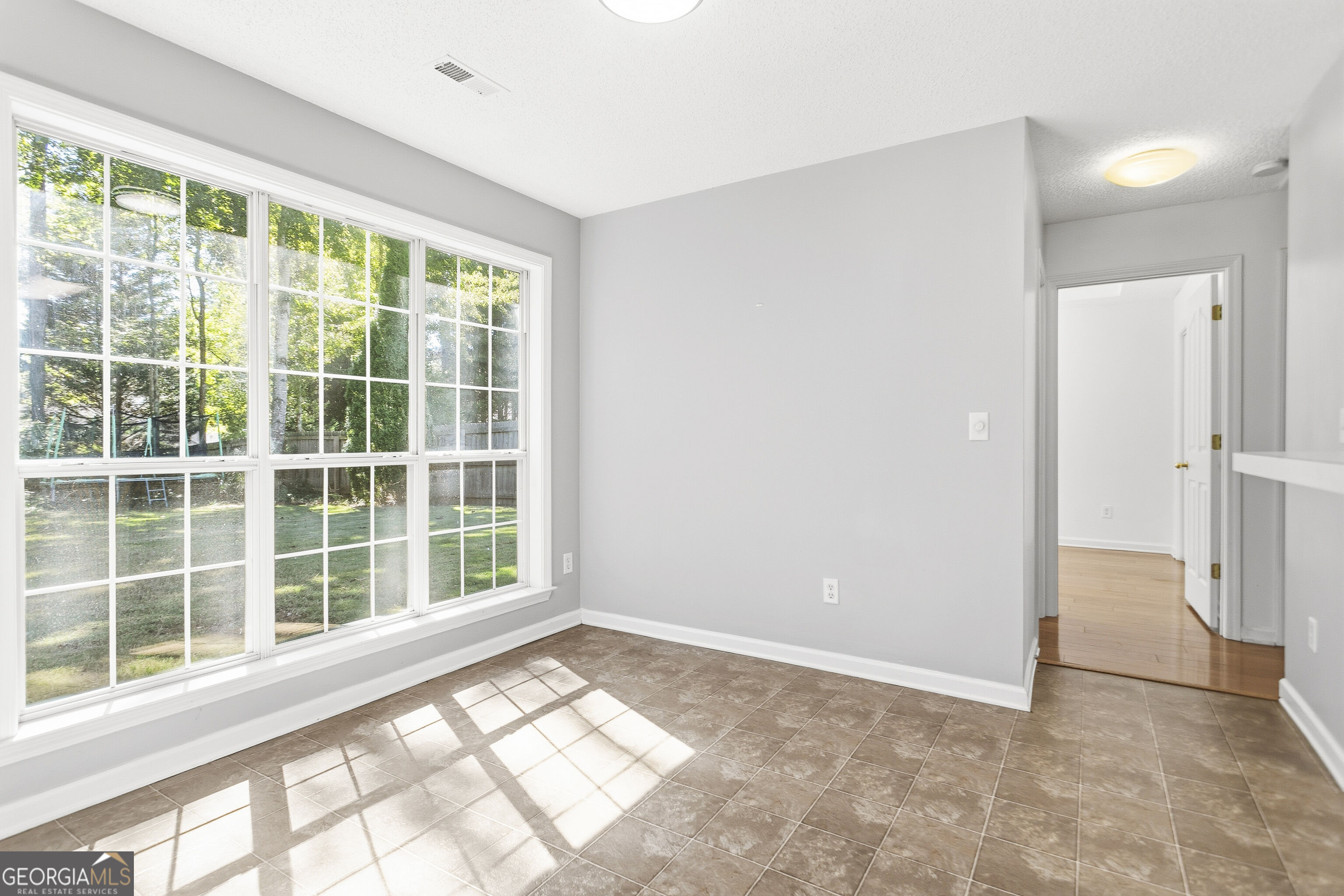 Vacant tiled room with large windows overlooking a green yard and a doorway to a hardwood-floored room.