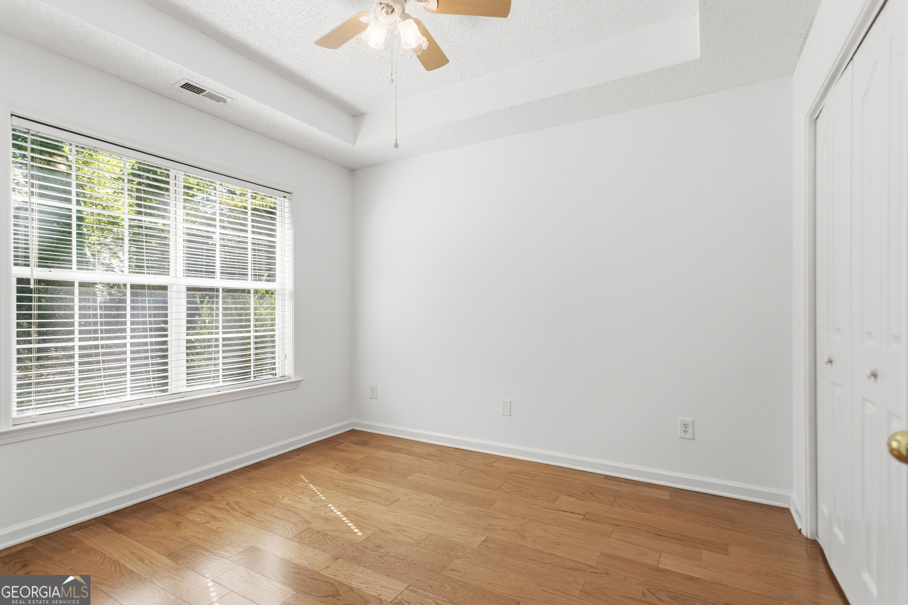 Empty bedroom with hardwood floors, a large window with blinds, and a ceiling fan.