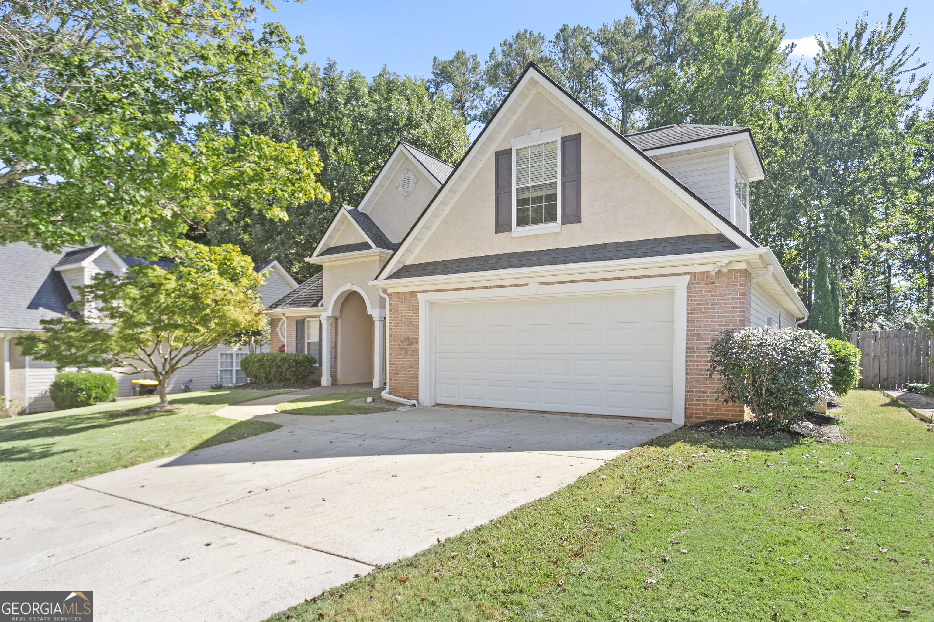 Two-story beige and brick home with prominent gables, a two-car garage, and a tree-lined lawn.