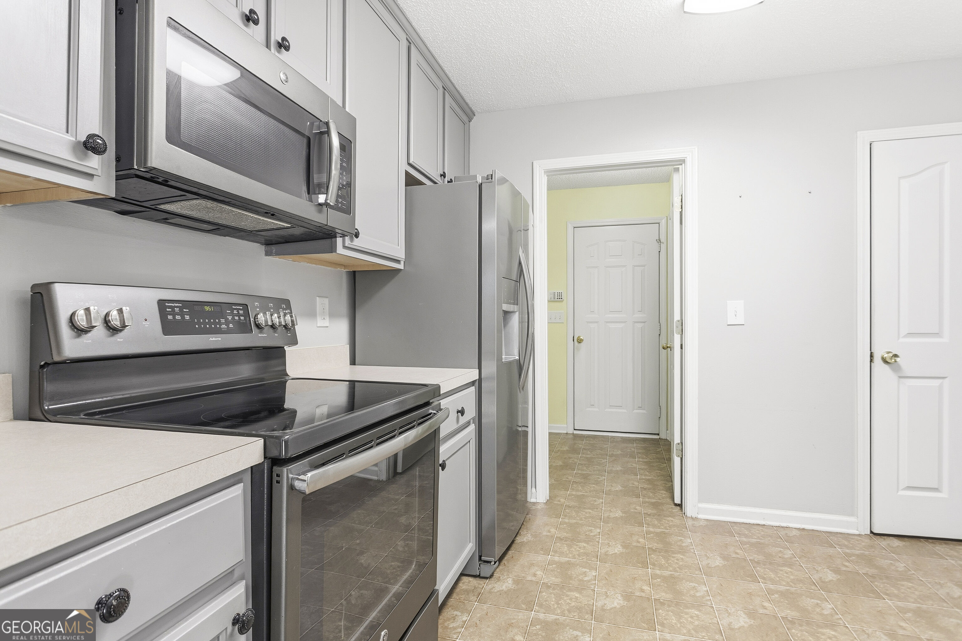 Kitchen with white cabinets, stainless steel appliances, and tan tile flooring leading to a hallway.