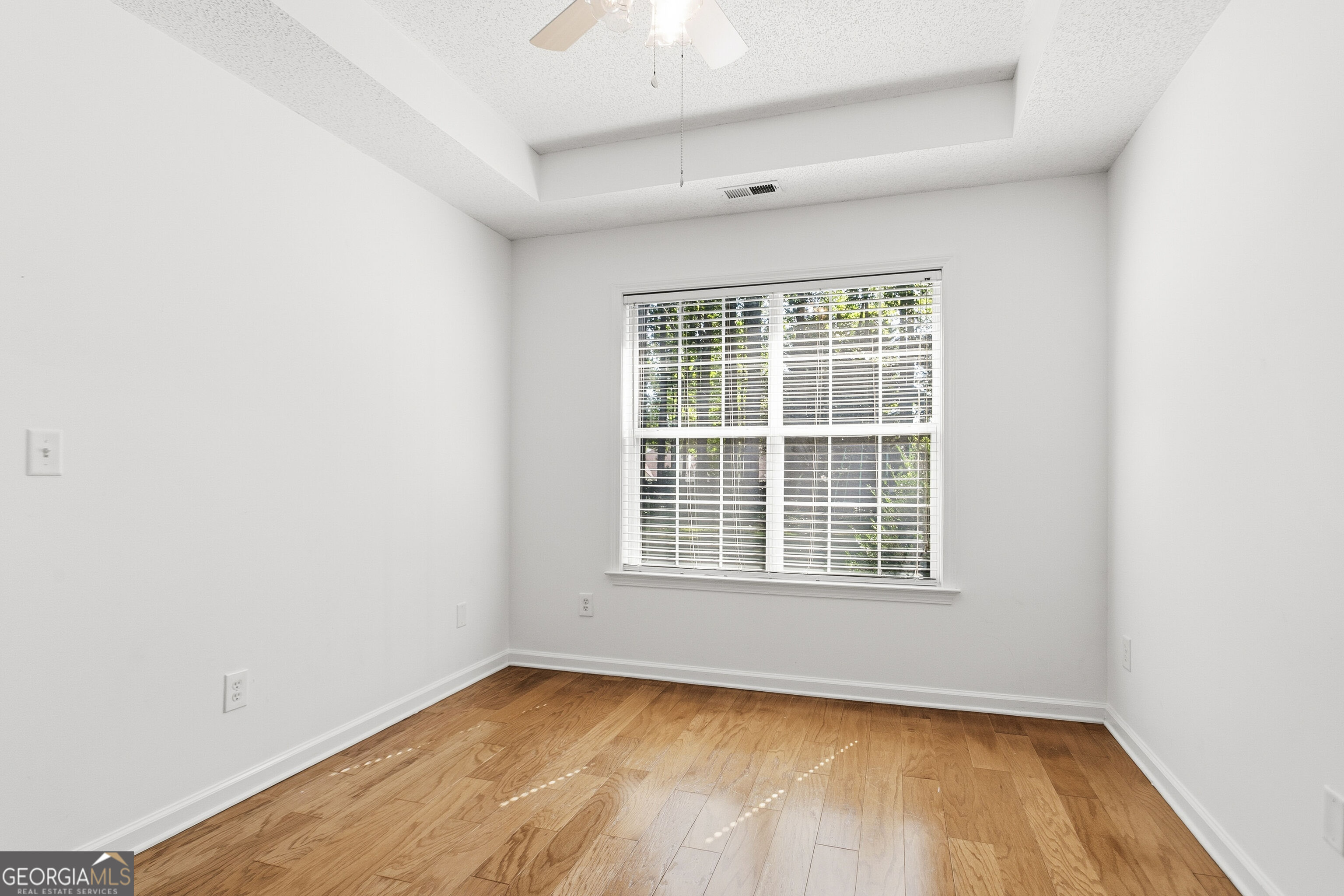 Empty room with hardwood floors, a raised ceiling, and a wide window with blinds.