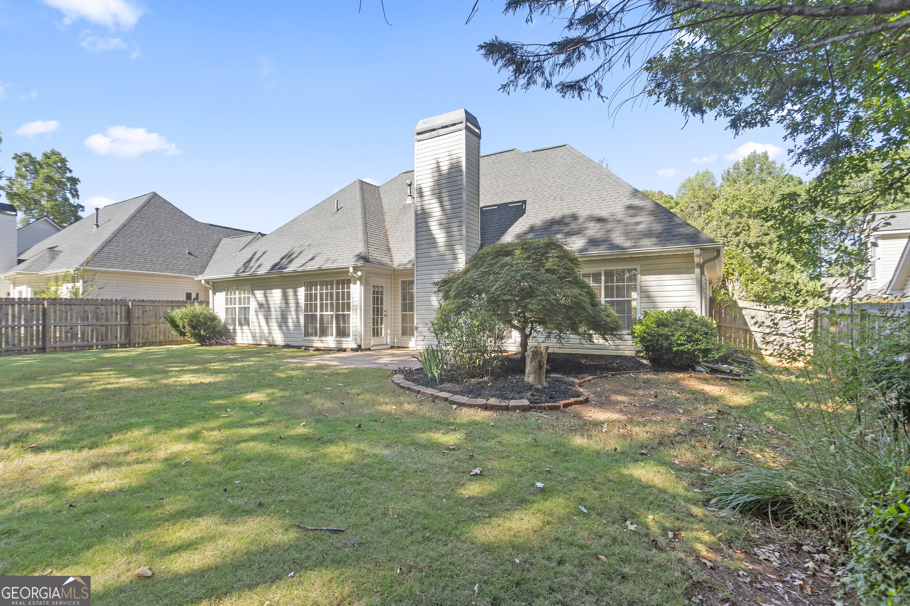 Rear of a gray-sided house with a tall chimney, fenced lawn, and landscaped planting bed.
