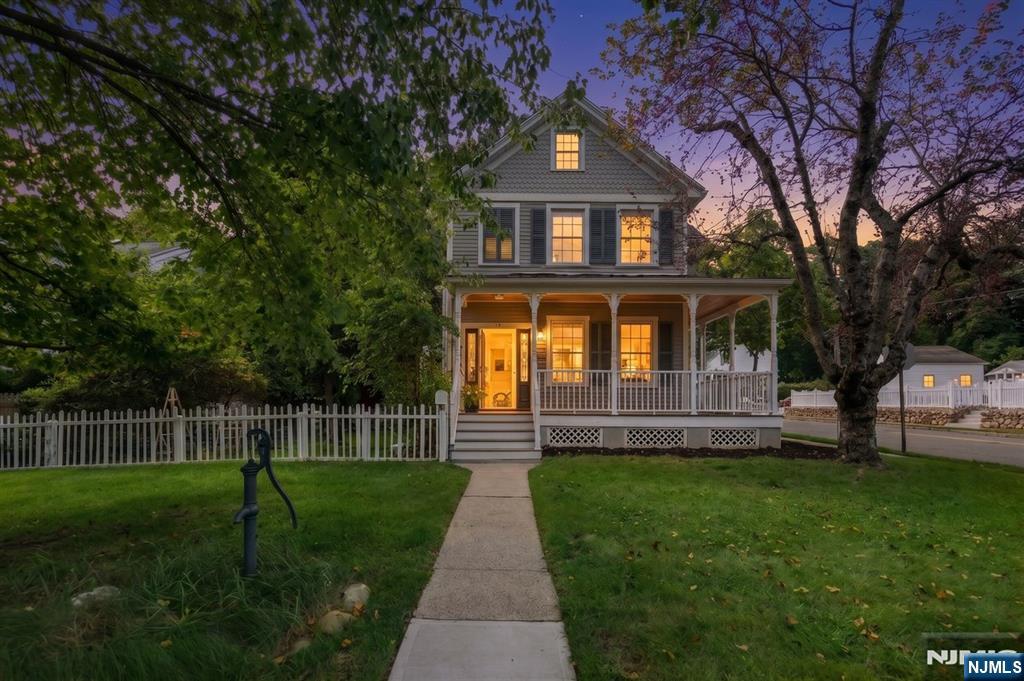 Two-story home with a covered porch, white picket fence, and illuminated windows at dusk.