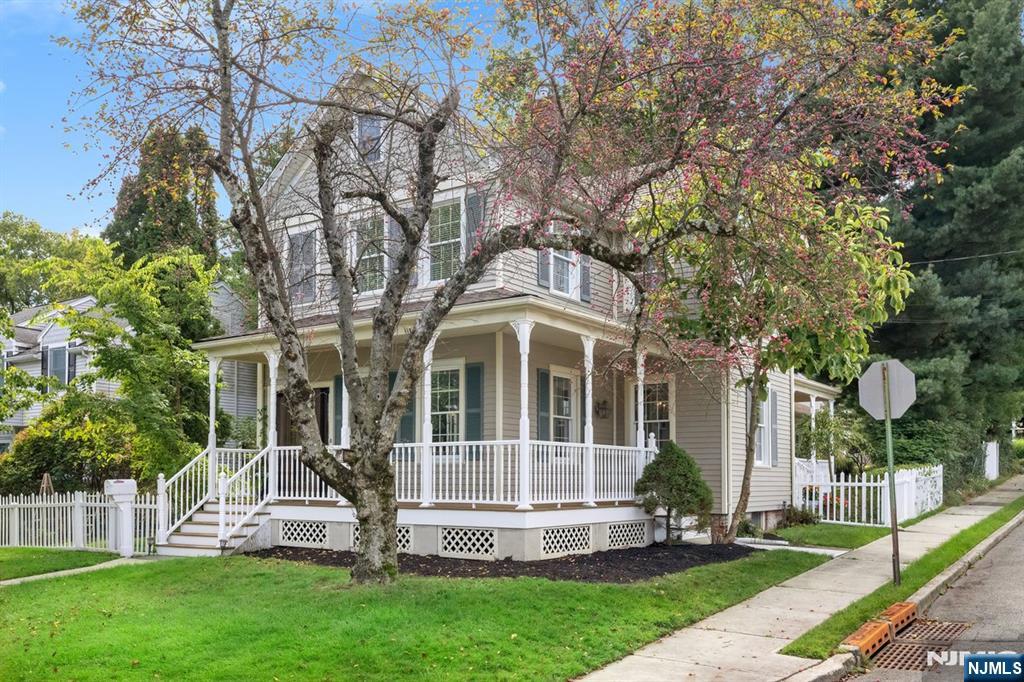 Two-story home with a covered porch and white railings, partly shaded by a flowering tree.