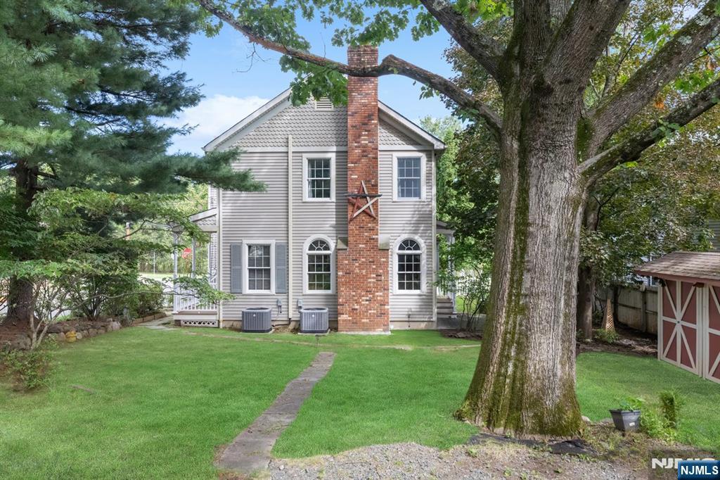 Rear of a gray two-story house with a brick chimney, broad lawn, mature trees, and a small shed.
