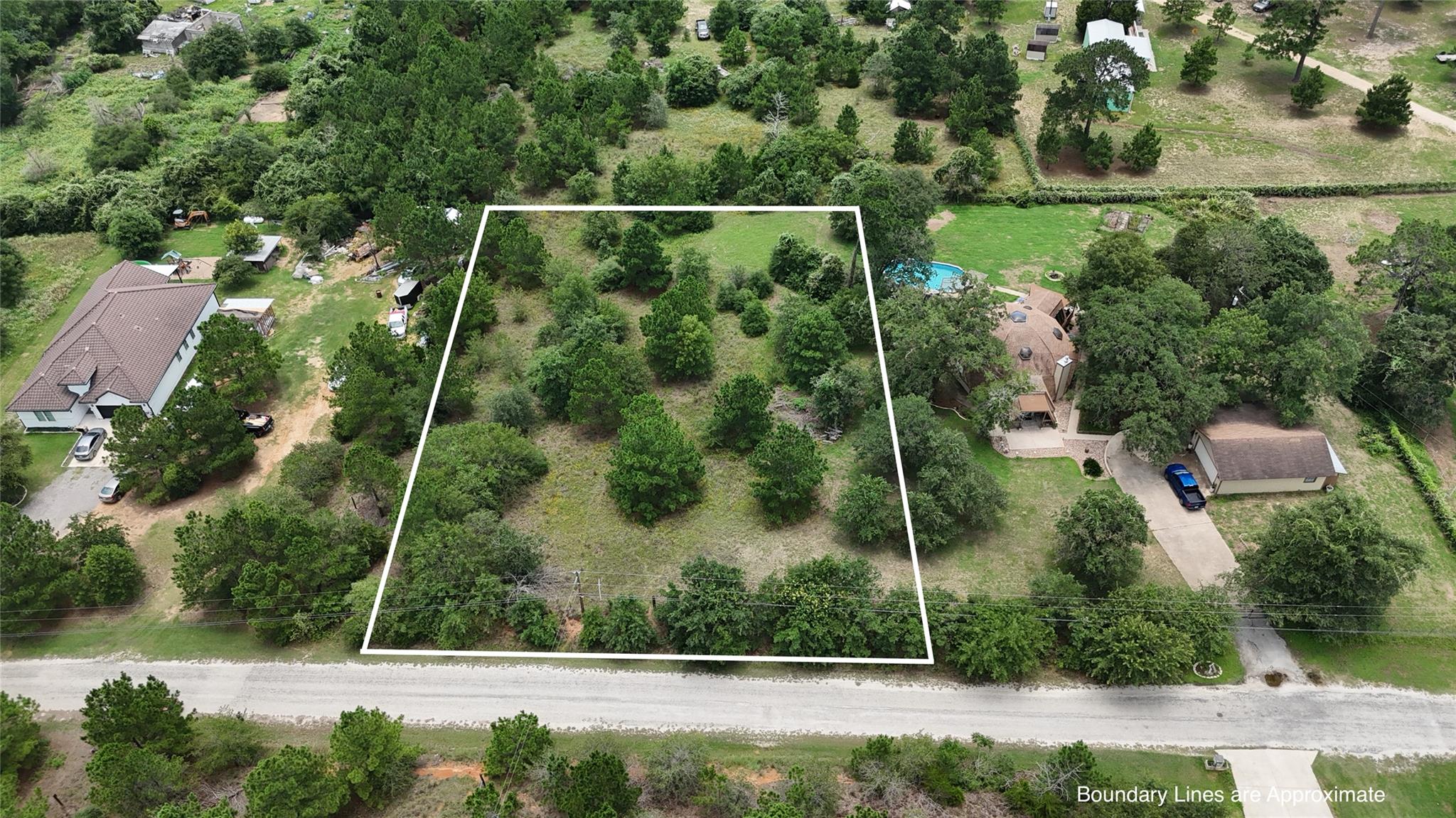 Aerial view of a wooded vacant lot outlined in white beside a road and neighboring homes.