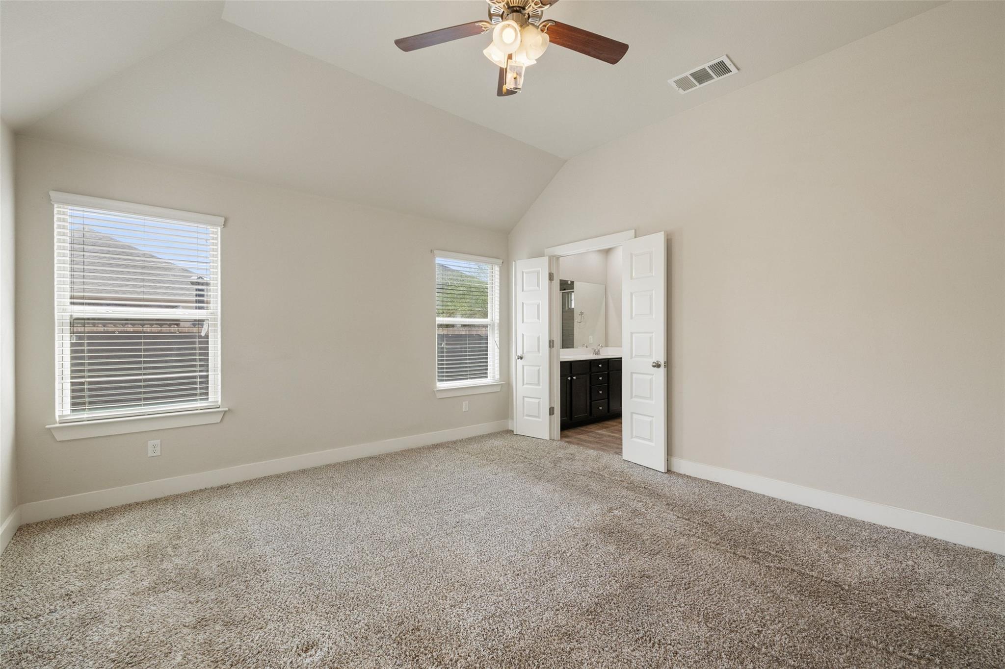 Empty carpeted bedroom with a ceiling fan, two windows, and open doors to a bathroom.